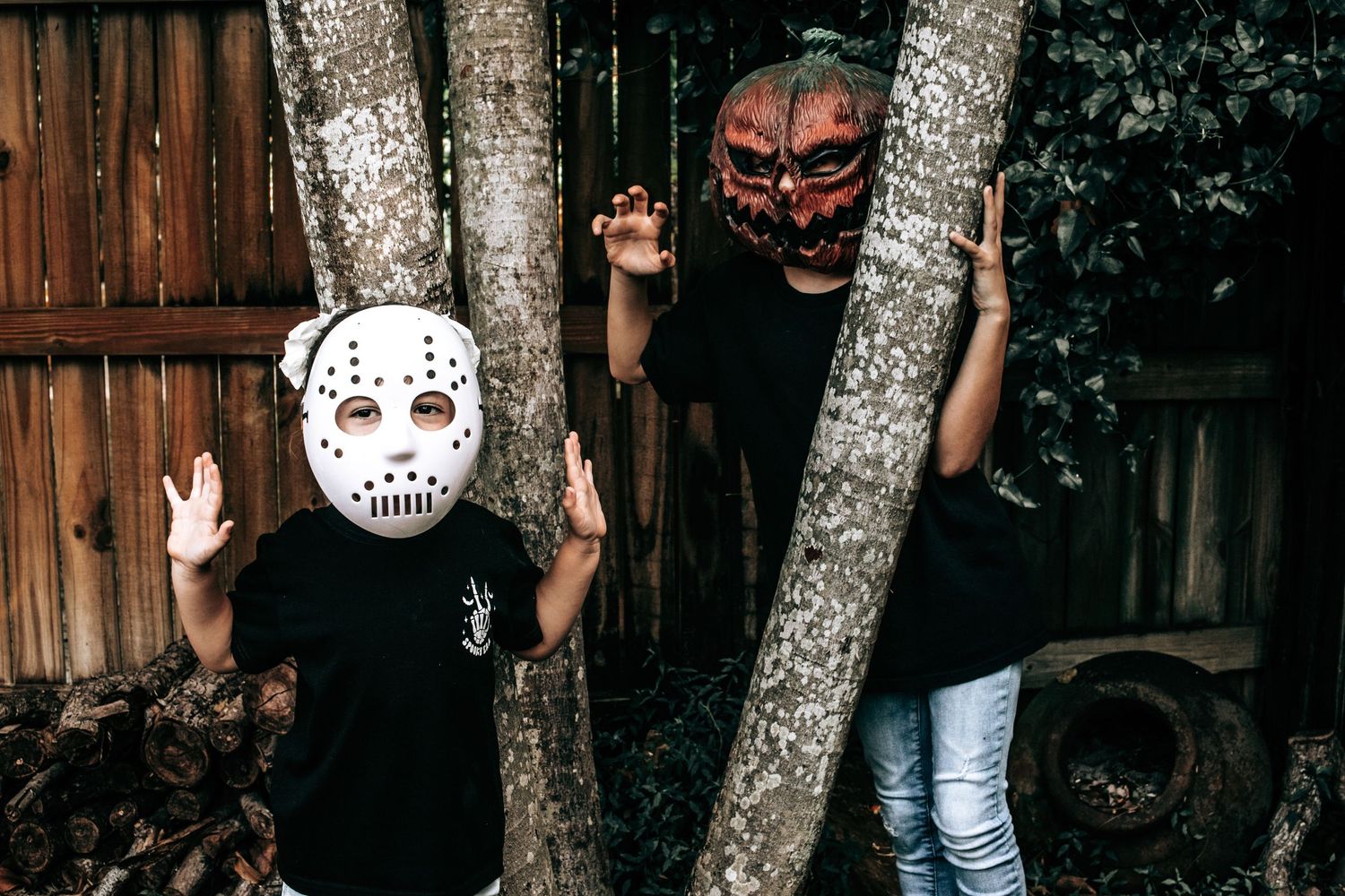 Two people in scary masks peek around tree trunks in Halloween setting.