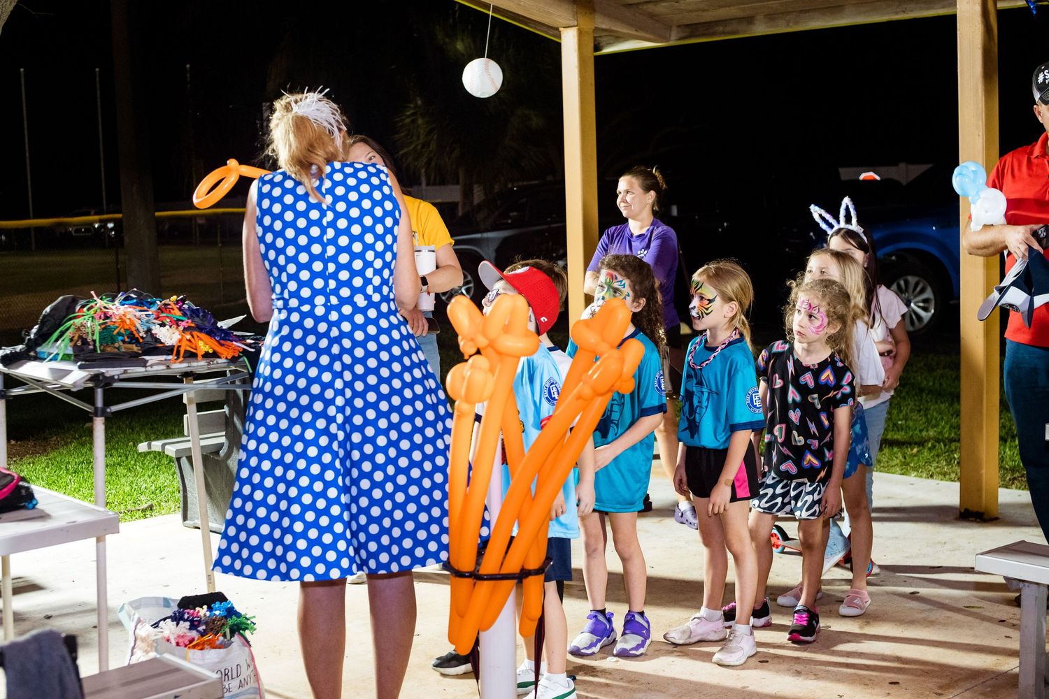 Children line up at an outdoor event while an adult in a polka dot dress leads an activity.