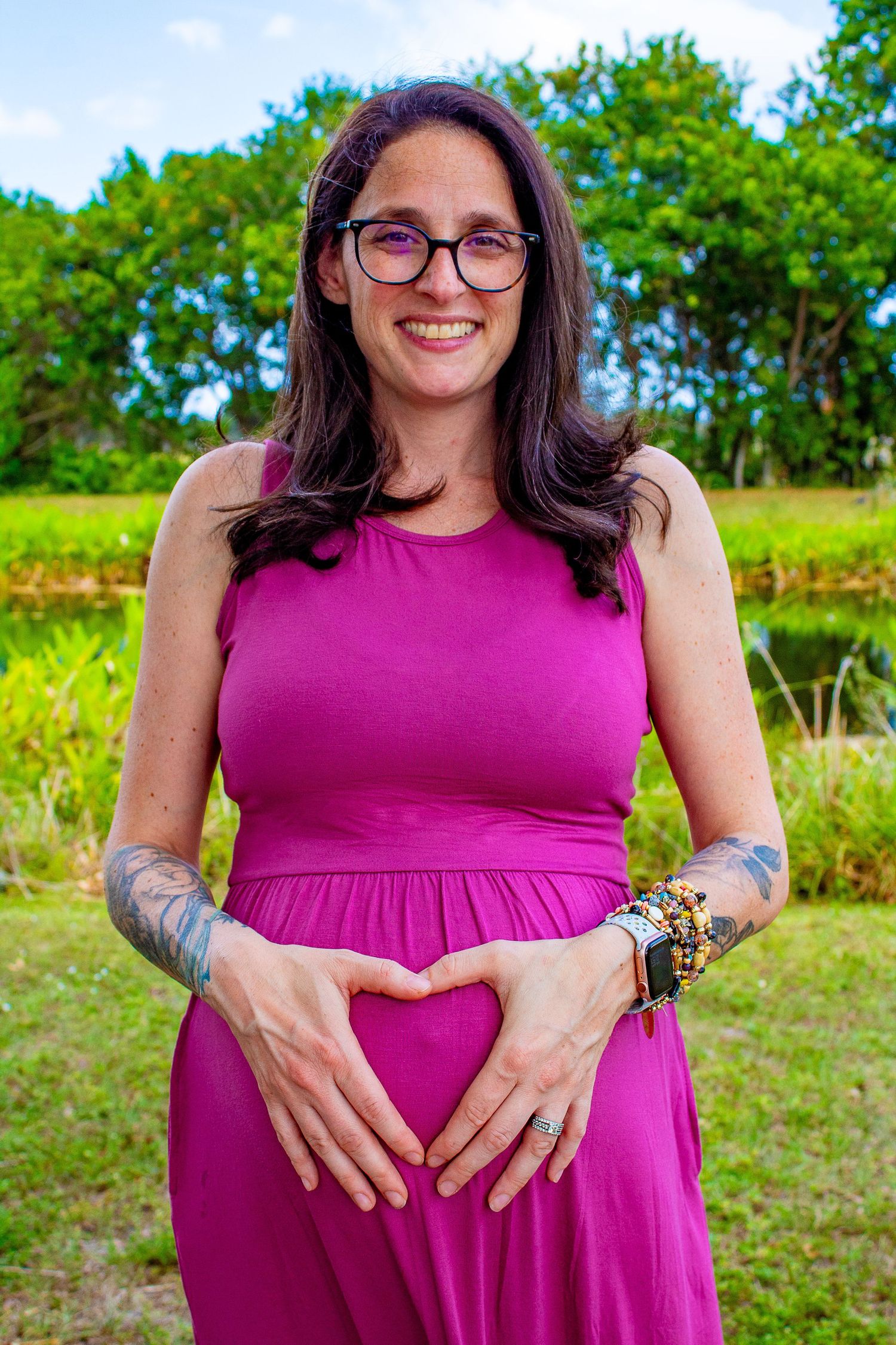 A person in a magenta dress stands in front of lush greenery outdoors.