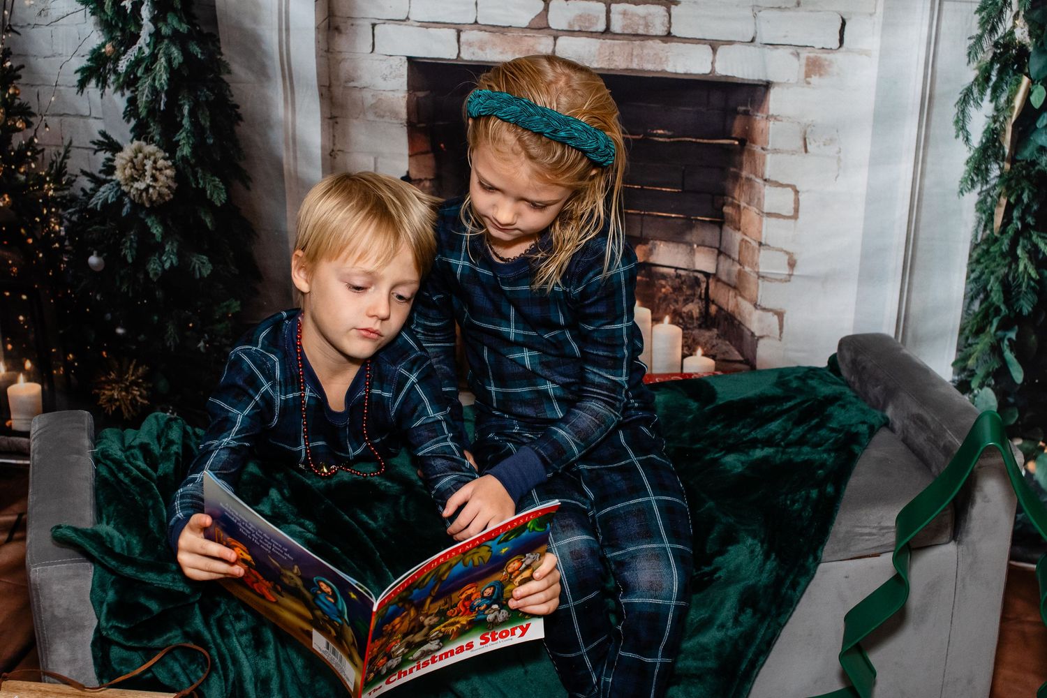 Two children in plaid pajamas read a book together on a velvet couch by a fireplace.