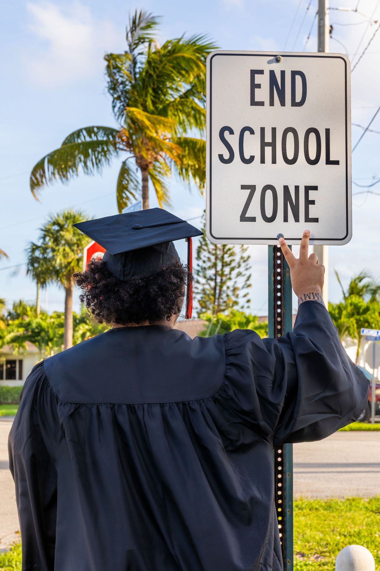 Graduate in cap and gown touching an End School Zone sign, with palm trees in the background.