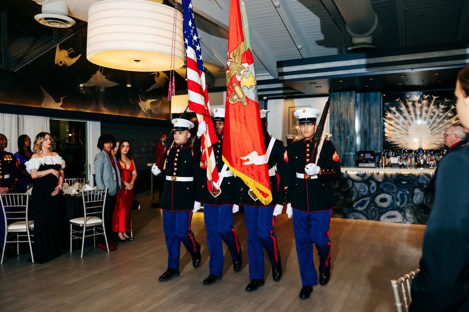 Military color guard in dress uniforms carrying American and Marine Corps flags during formal ceremony presentation.