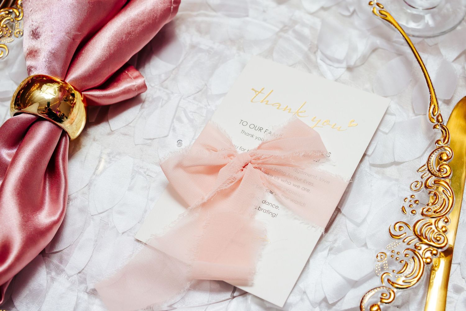 Elegant table setting with pink silk ribbons and gold ornate details on white background.