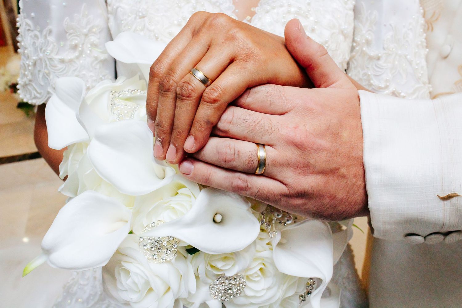 Close-up of bride and groom's hands with wedding rings resting on white calla lily bouquet.