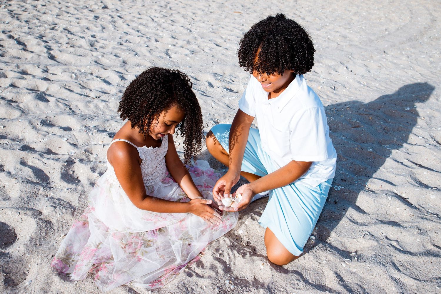 Two people sit together on a white sandy beach while sharing a tender embrace in the bright sunlight.