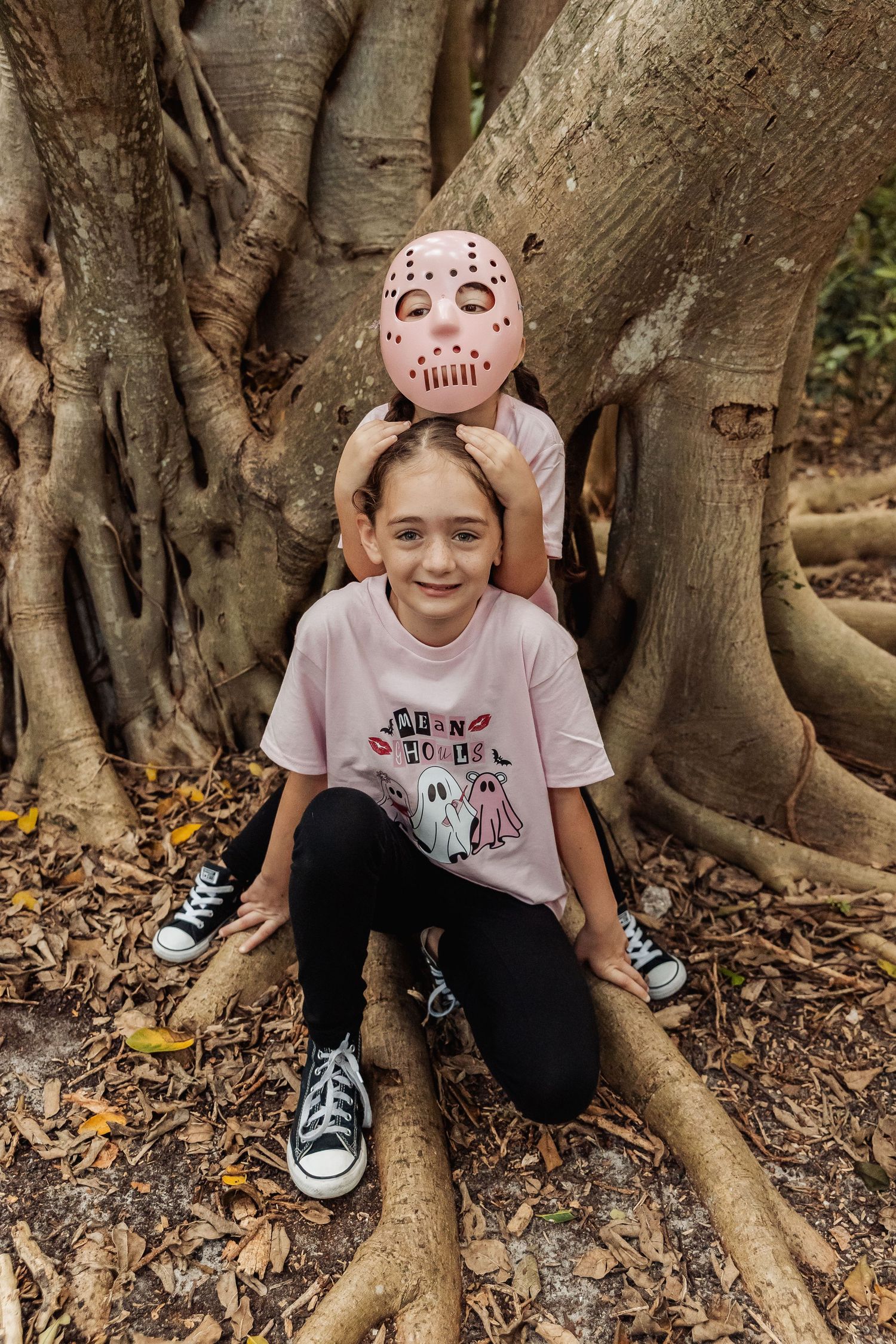 Child wearing face paint crouches by large tree roots in a forest setting with fallen leaves on the ground.