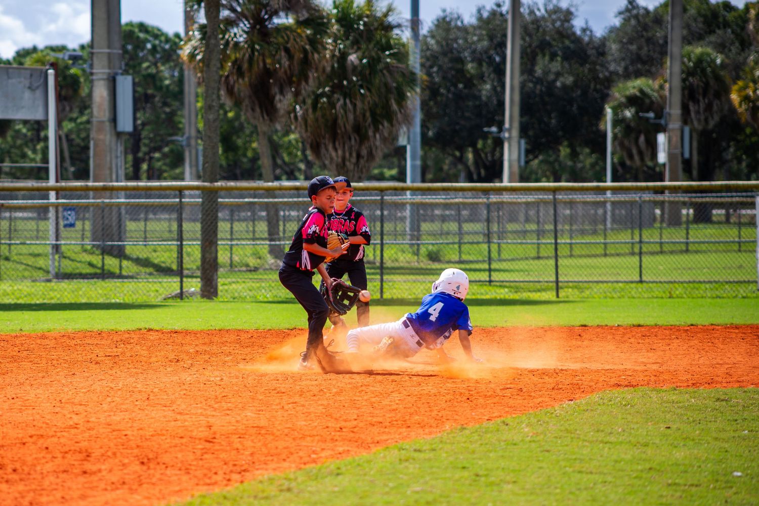 Baseball player in red jersey attempts to tag runner sliding into base on orange clay infield during game.