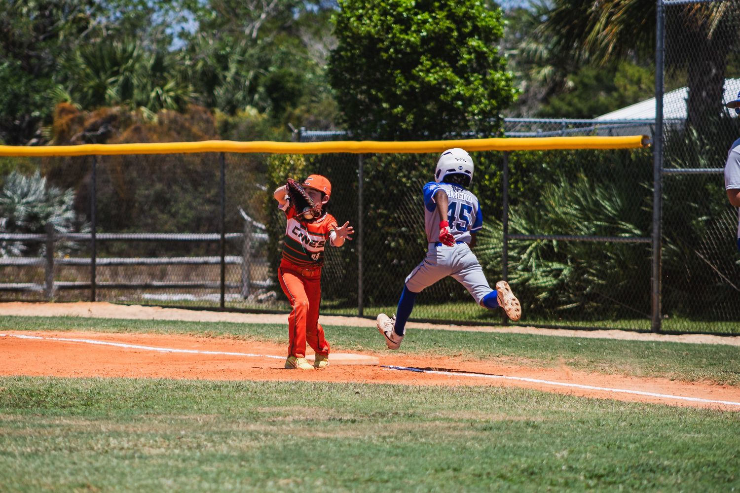 Youth baseball players compete on a sunny day as a runner leaps towards first base while the baseman awaits the throw.