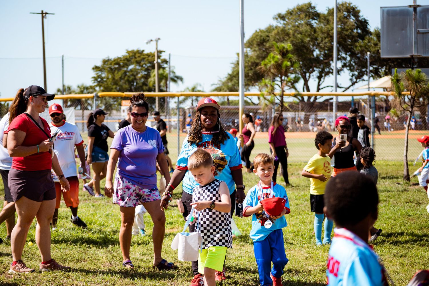 People gather outdoors at a community event in a park on a sunny day with trees in the background.