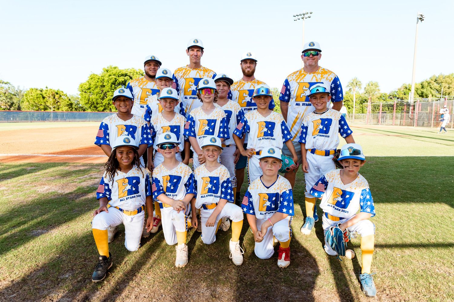 Youth baseball team wearing white and blue uniforms poses together on a sunny field for their team photo.