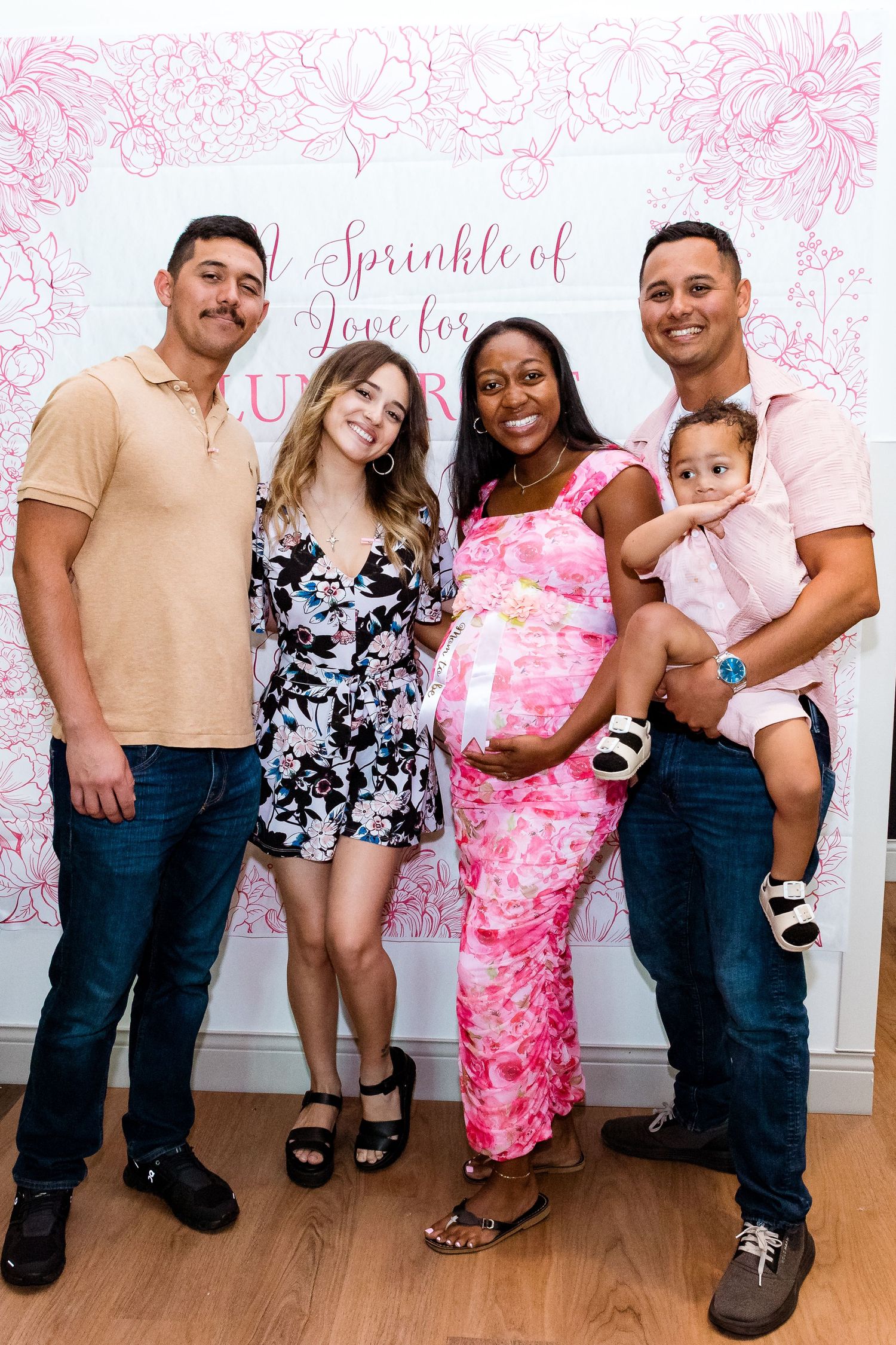Group posing together at a baby shower celebration with pink floral backdrop showing a pregnant woman in pink dress.