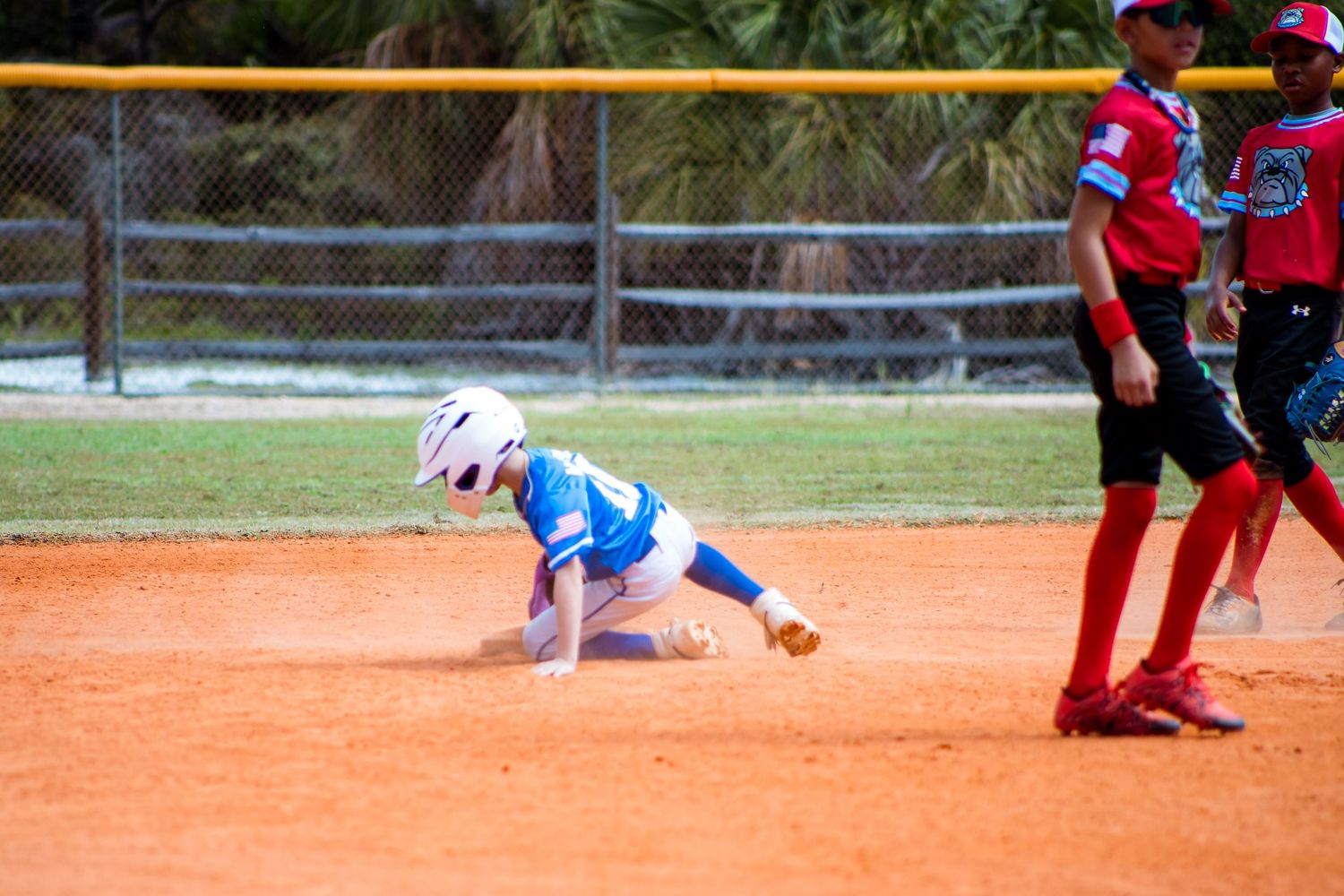 Softball players in red and white uniforms fielding during a game on a dirt infield.
