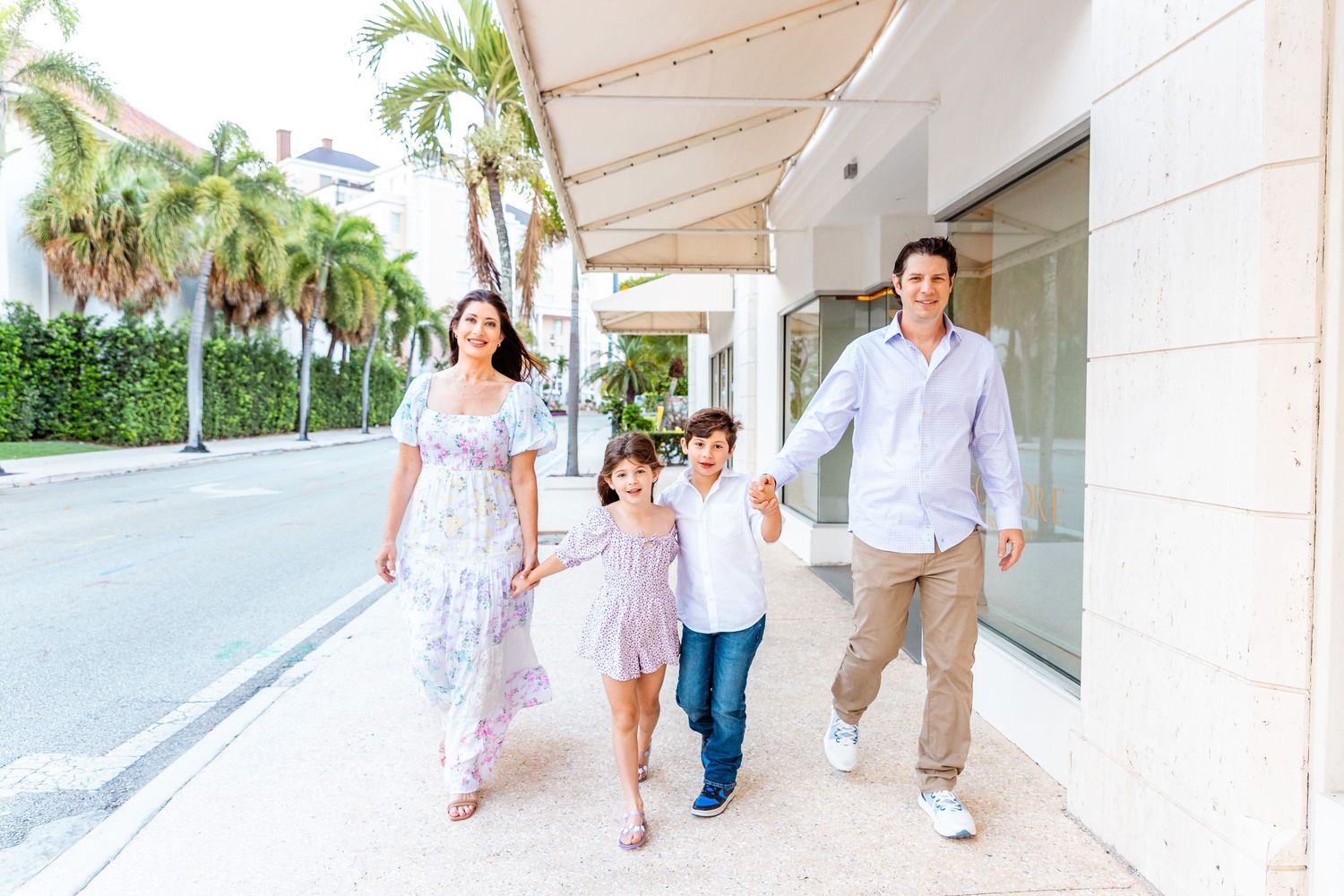 Happy family of four walking hand-in-hand on a sunny sidewalk lined with palm trees in a tropical town.