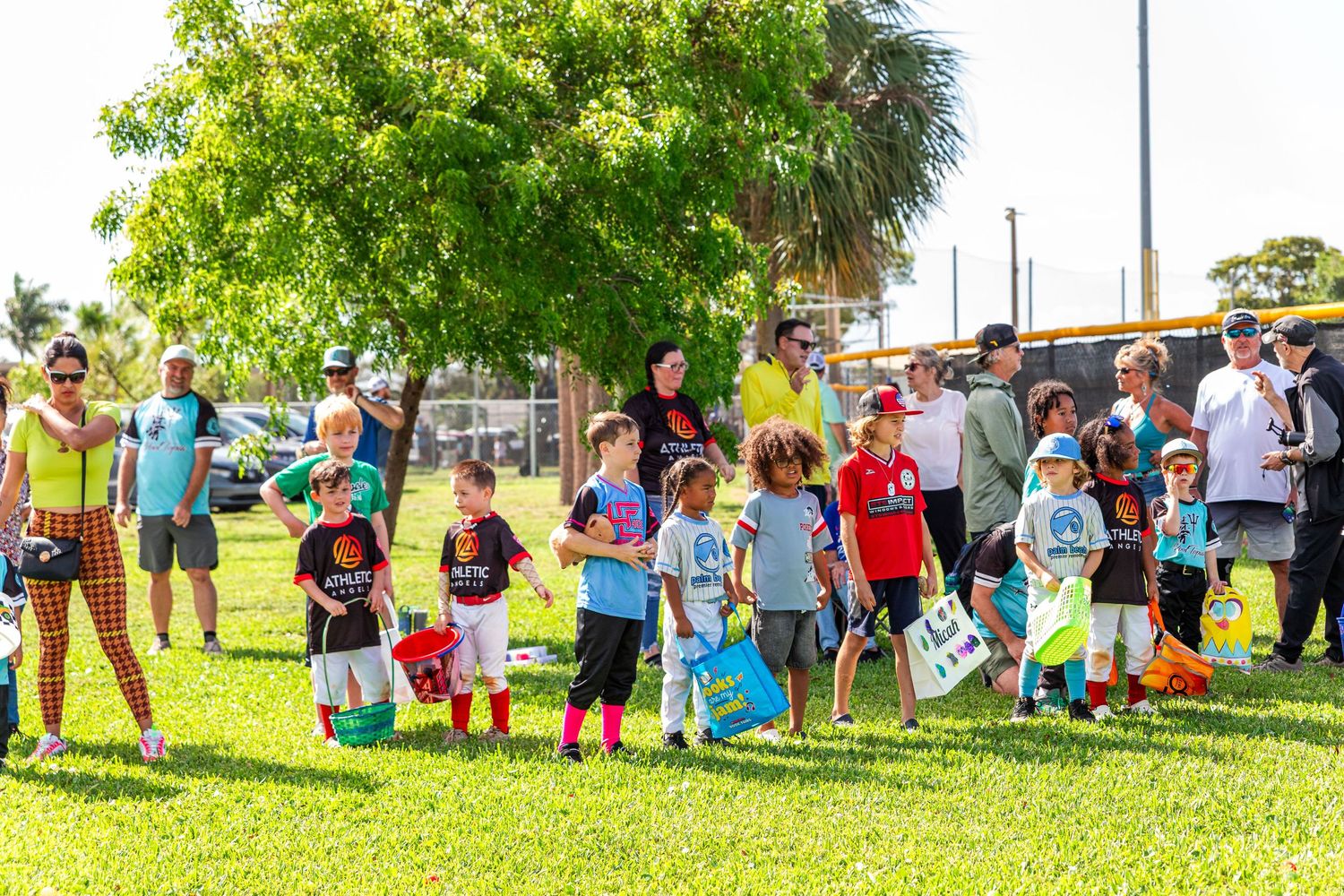 Children and adults gather on green grass under trees at an outdoor community event on a sunny day.