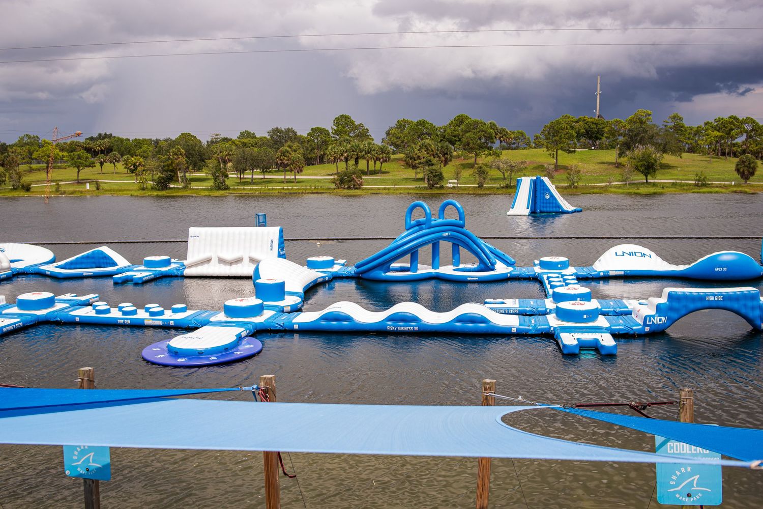 Large inflatable water park with blue and white floating obstacles and platforms set up on a lake under cloudy skies.
