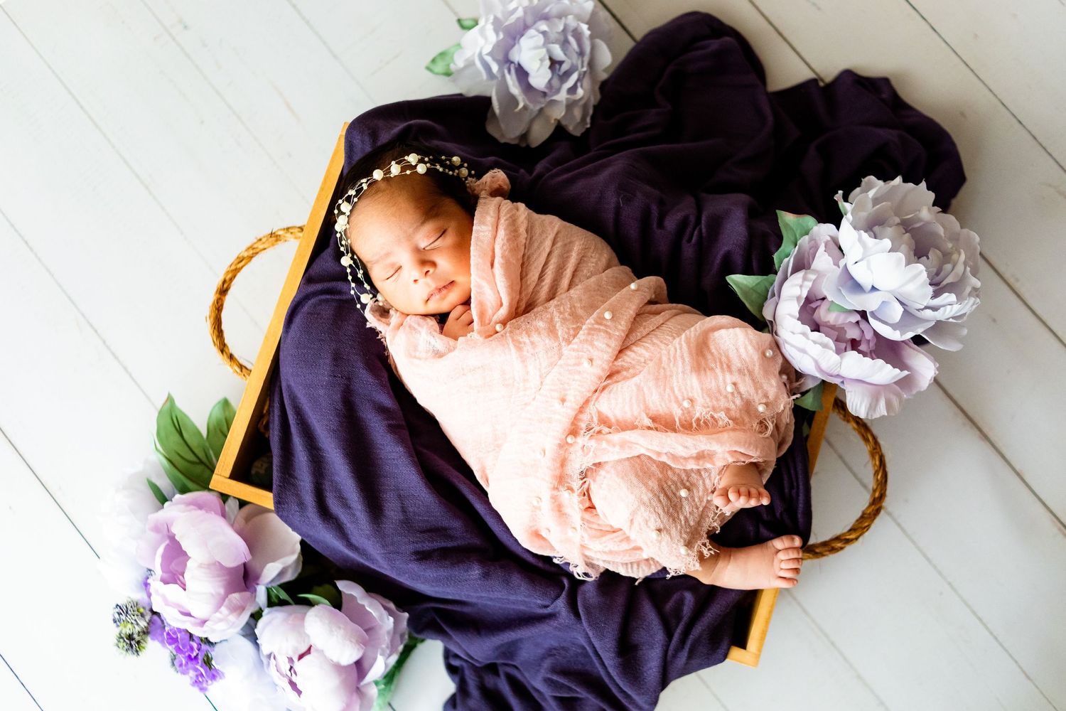 Newborn baby girl swaddled in pink lace wrap sleeping in a wicker basket with purple fabric and floral decorations.