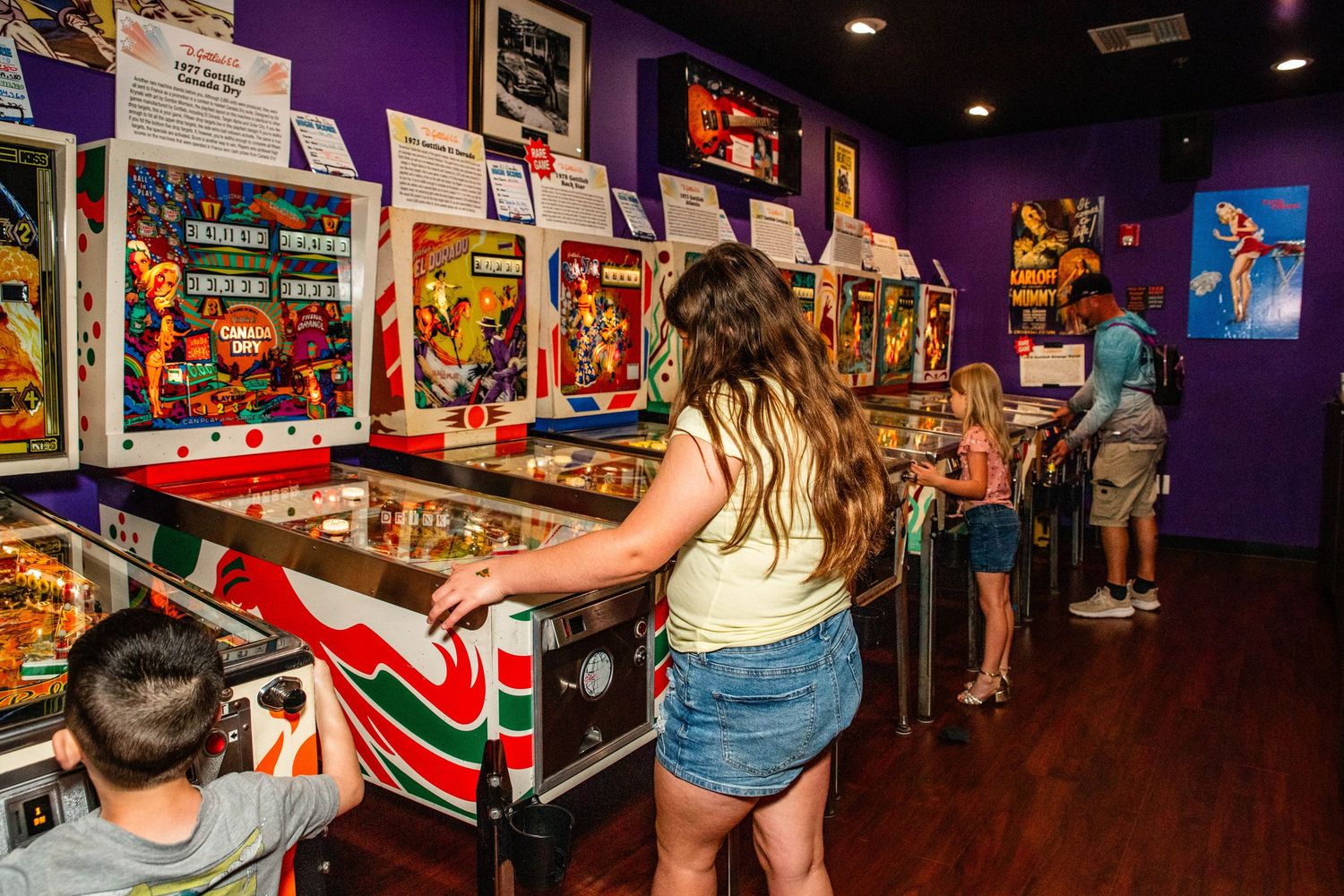 The image shows a young woman playing a vintage pinball machine in an arcade setting with colorful walls and decor.