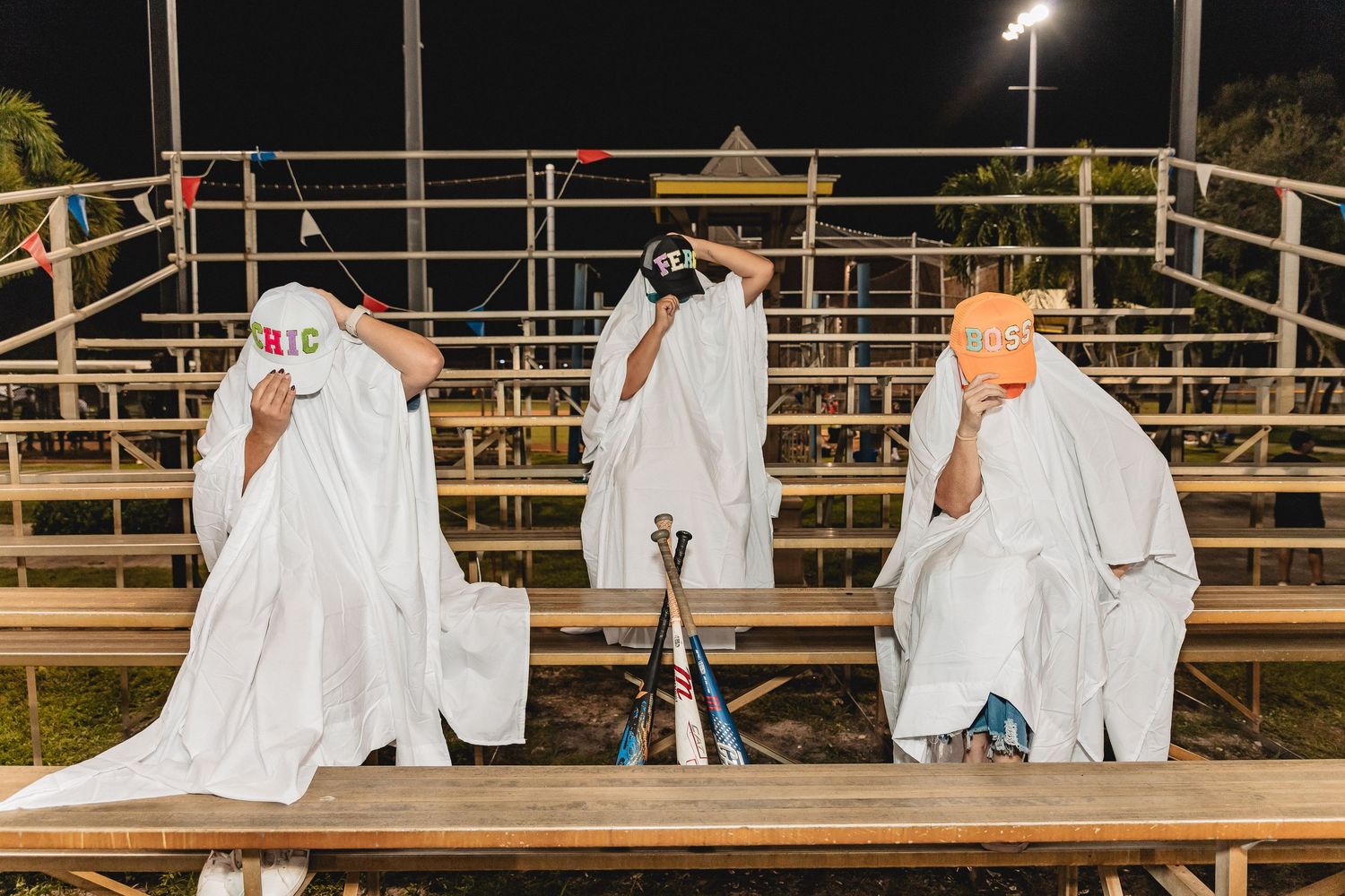 Row of figures in white ghost costumes stand on bleachers at a nighttime baseball field.
