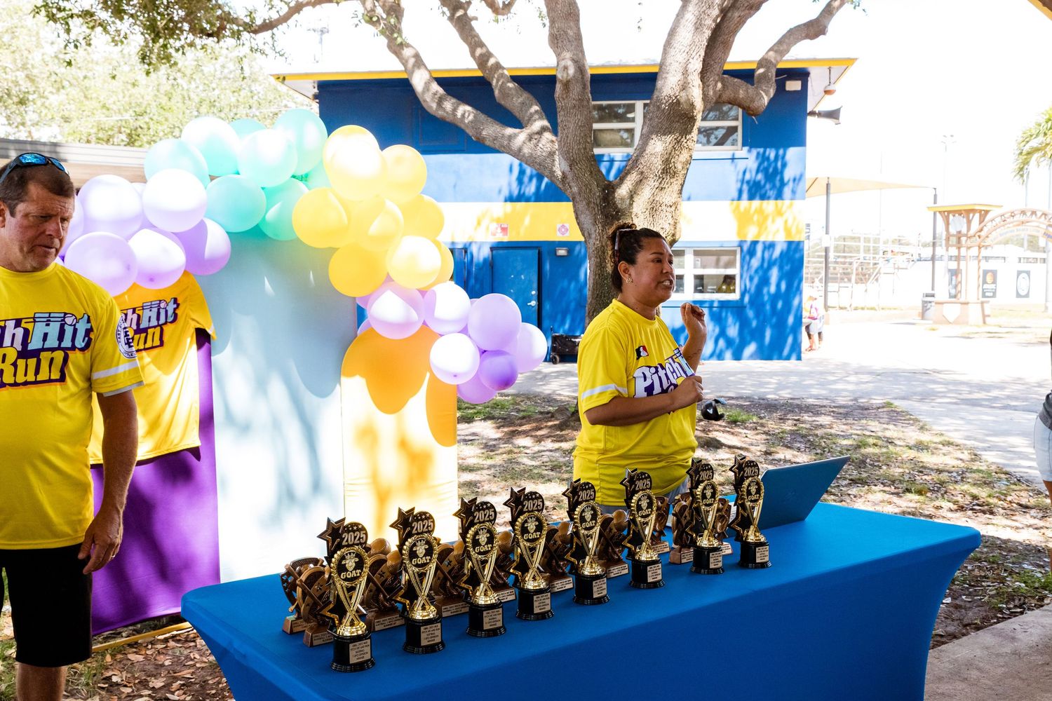 Display table with trophy awards and colorful balloon arch decorations at outdoor event with yellow-shirted staff.