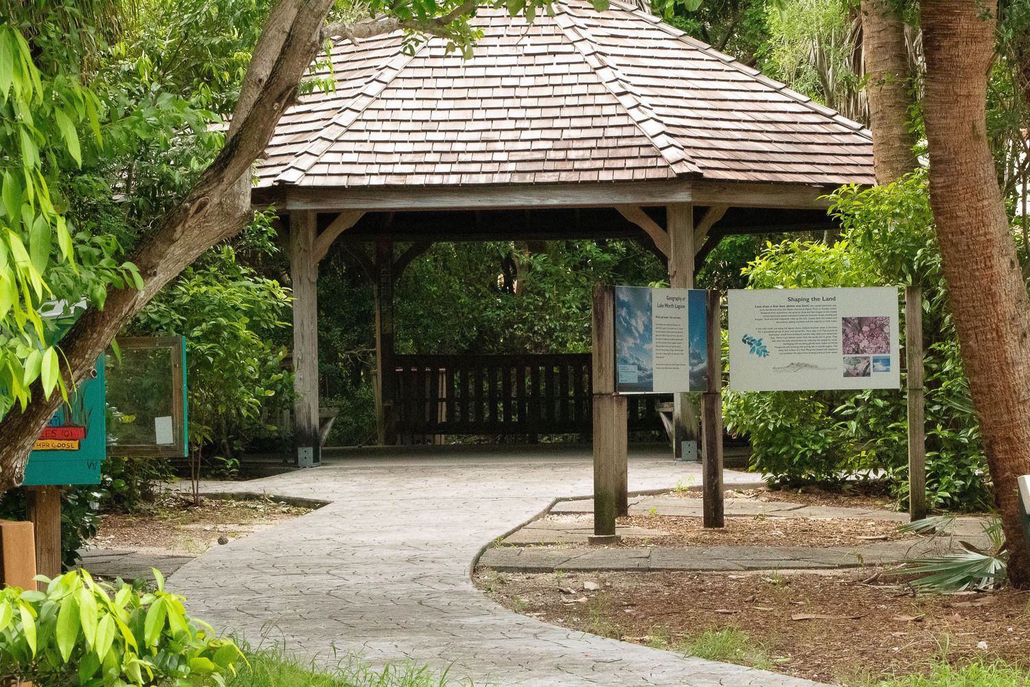 A wooden gazebo with a shingled roof stands along a curved pathway in a lush tropical garden setting.