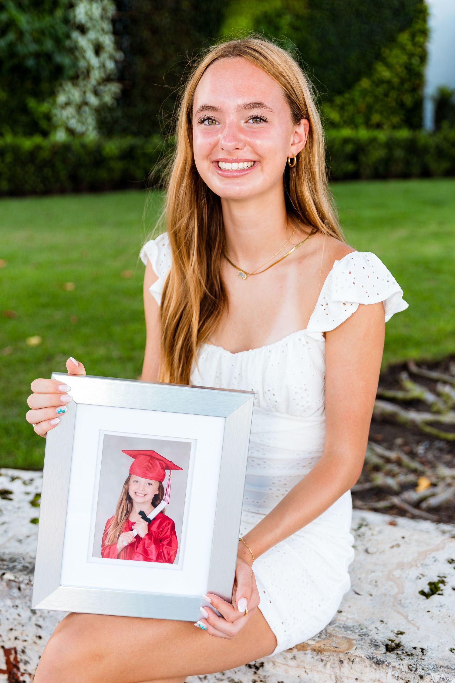 Young woman in white dress poses outdoors while holding a framed graduation photo on a sunny day.