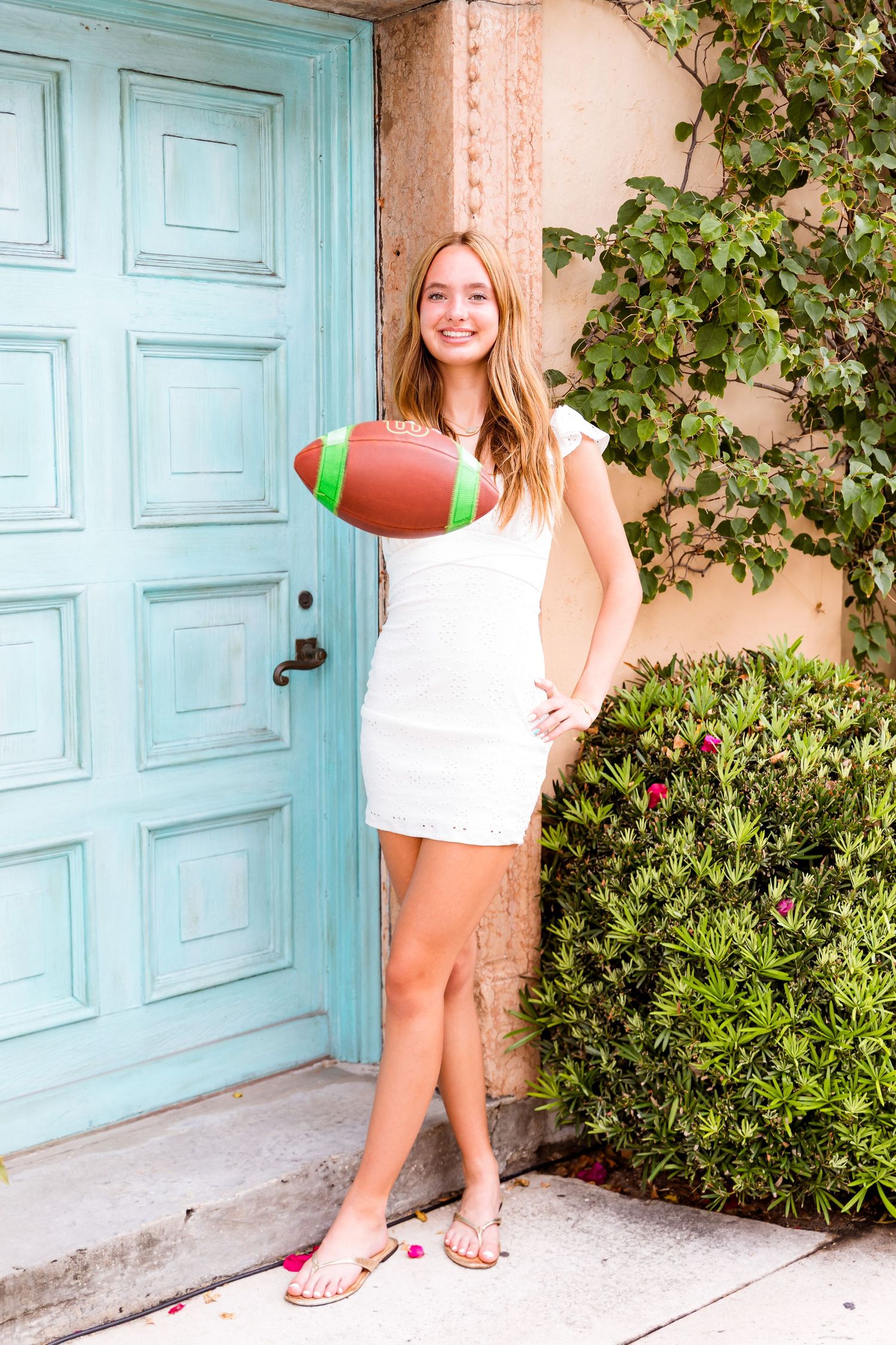 A cheerful figure in a white dress poses by a turquoise door holding a football in front of a vine-covered wall.