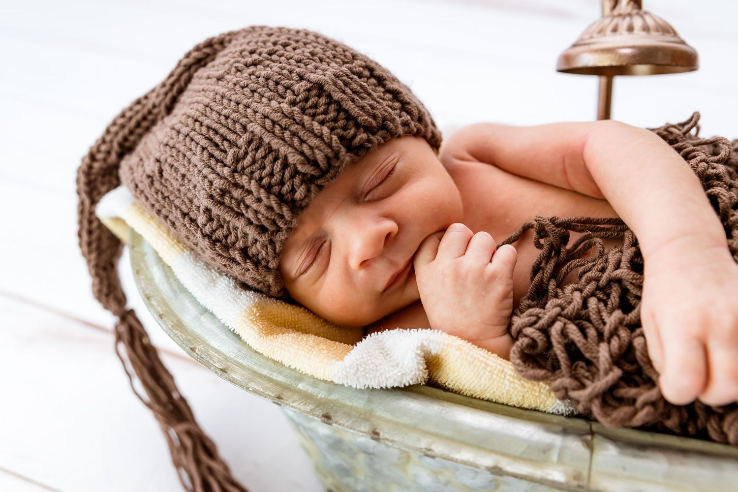 Sleeping newborn wearing brown knit hat rests peacefully in vintage basket with soft blankets.