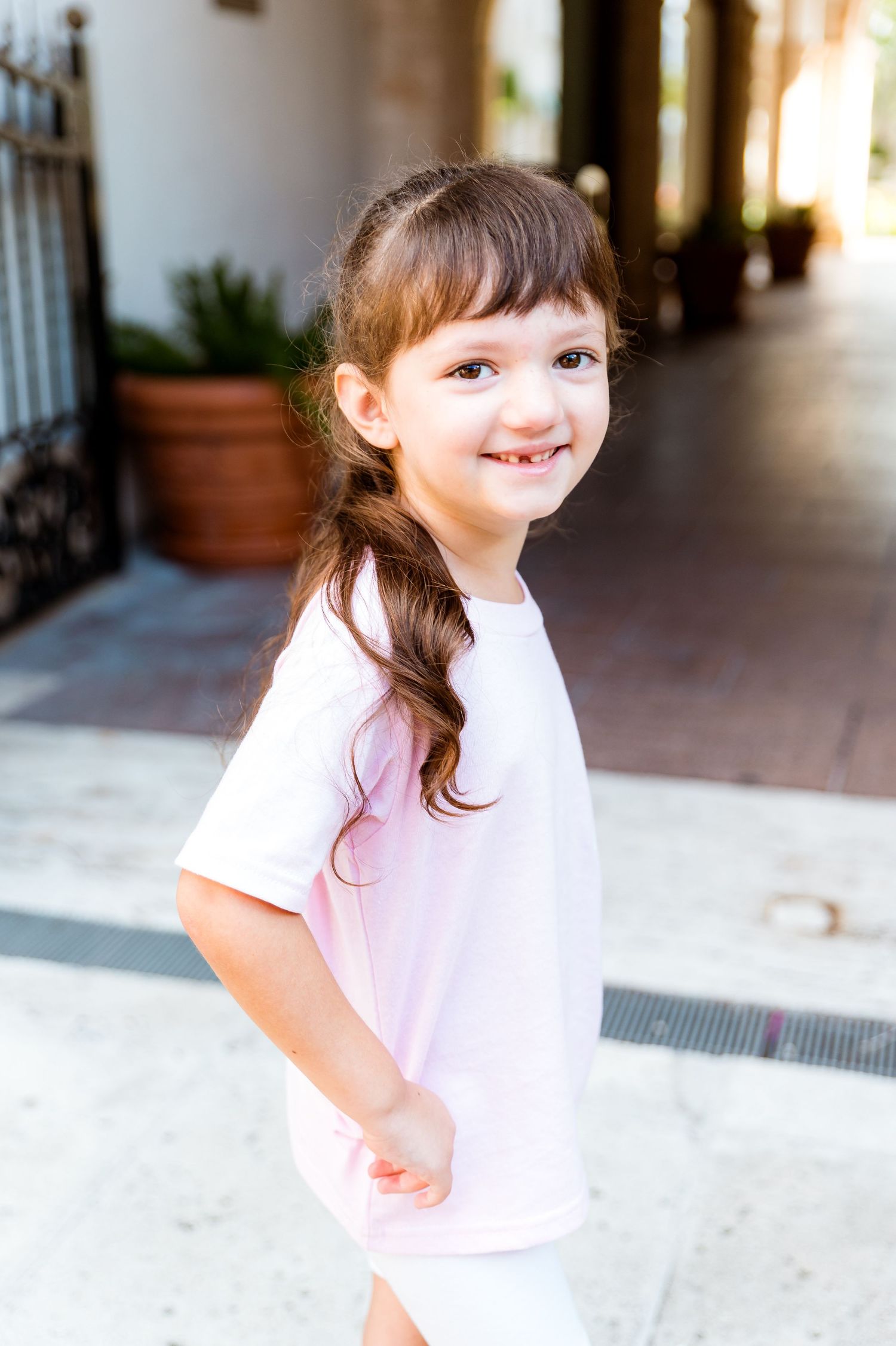 A young child in a white t-shirt with brown braided hair standing in a covered outdoor walkway.