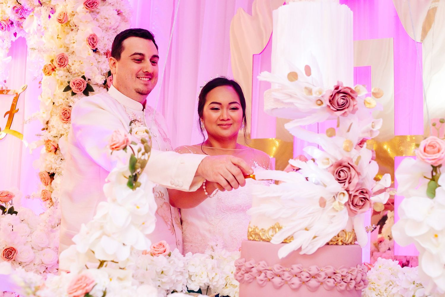 Couple cutting their elegant white wedding cake surrounded by pink and floral decorations.