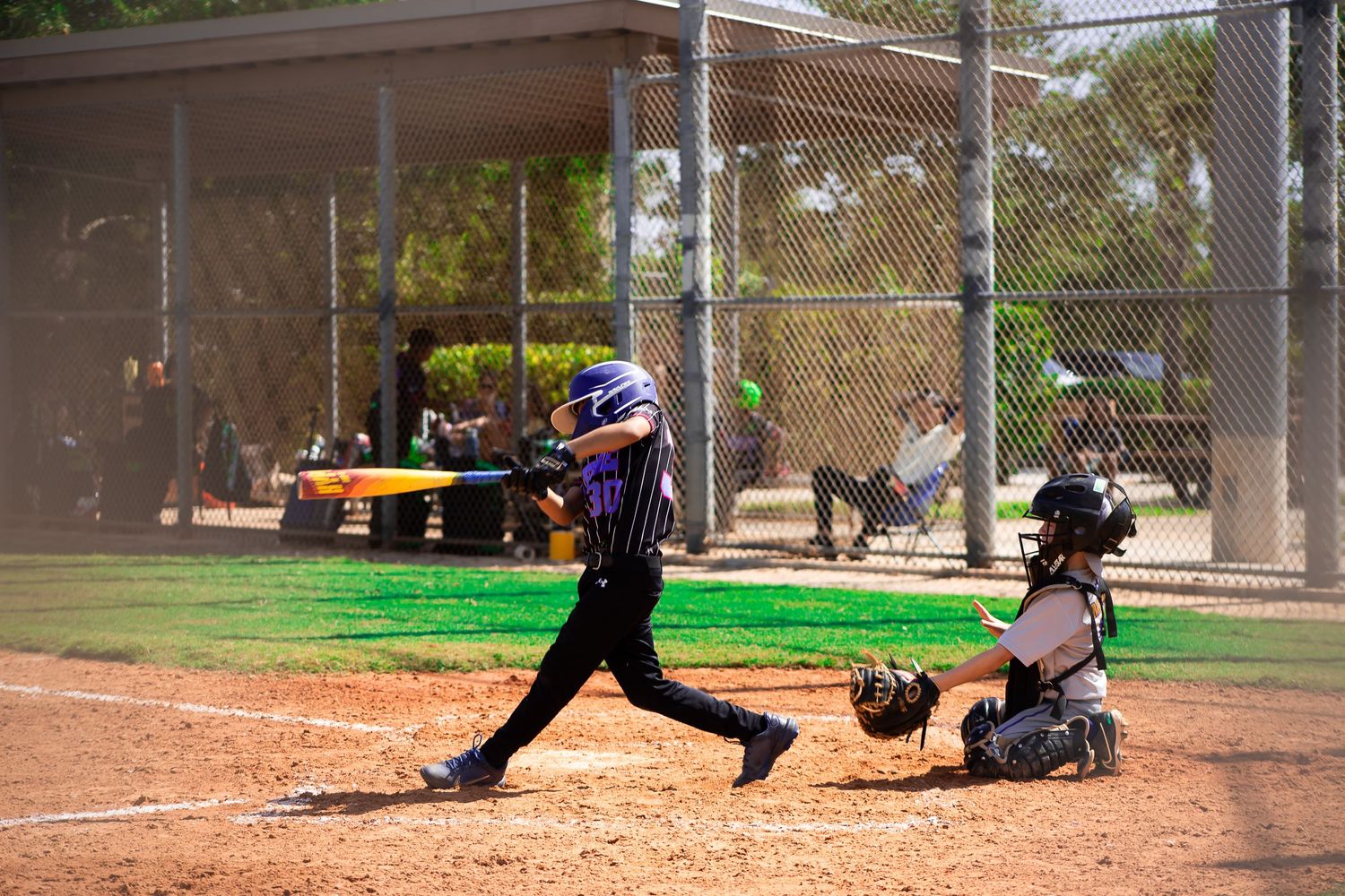 Youth baseball batter swings at pitch while catcher crouches behind home plate during game on sunny day.