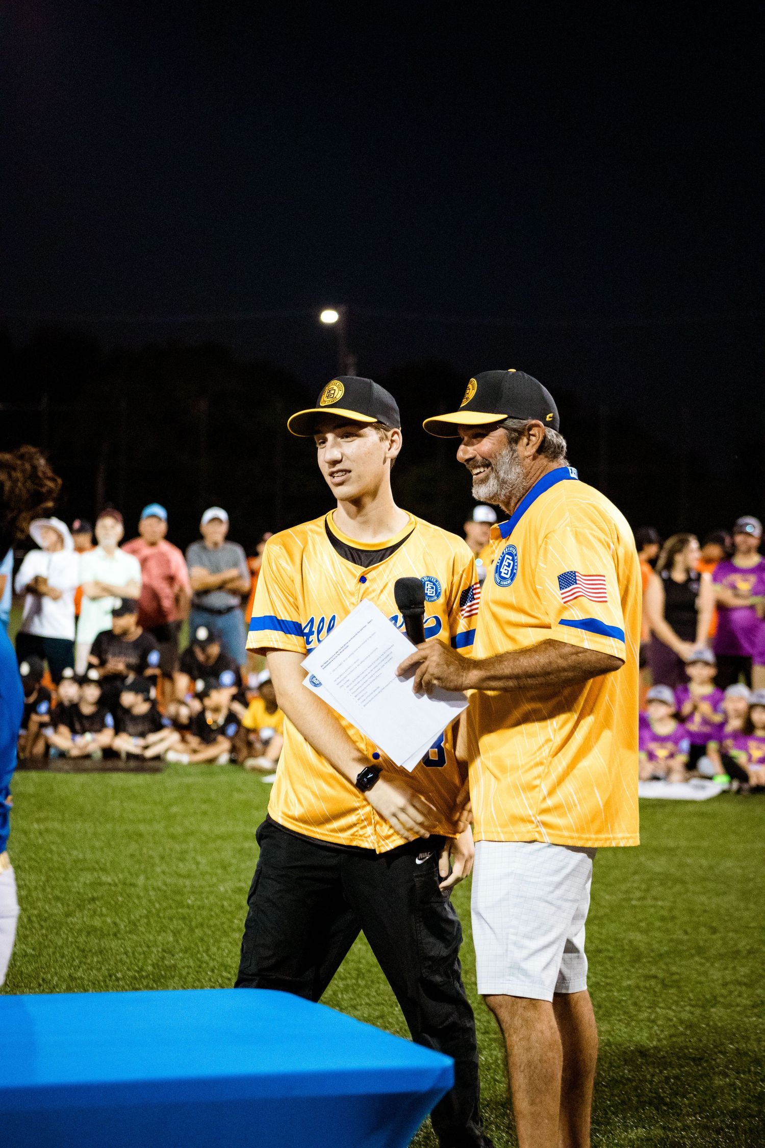 Two baseball coaches in yellow jerseys review game notes under stadium lights during a nighttime event.