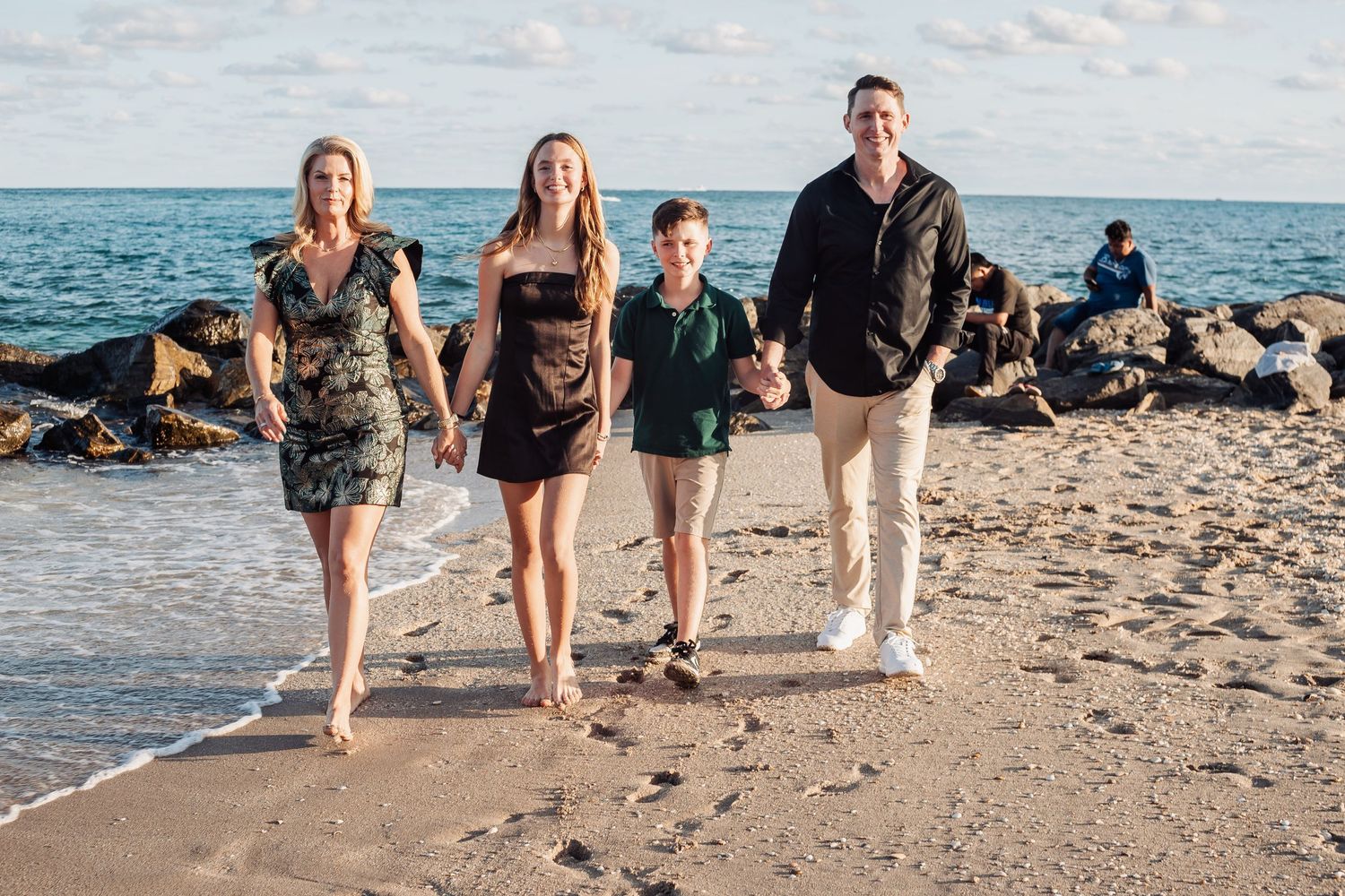 Family of four walking together on a beach at sunset with rocky coastline in the background.