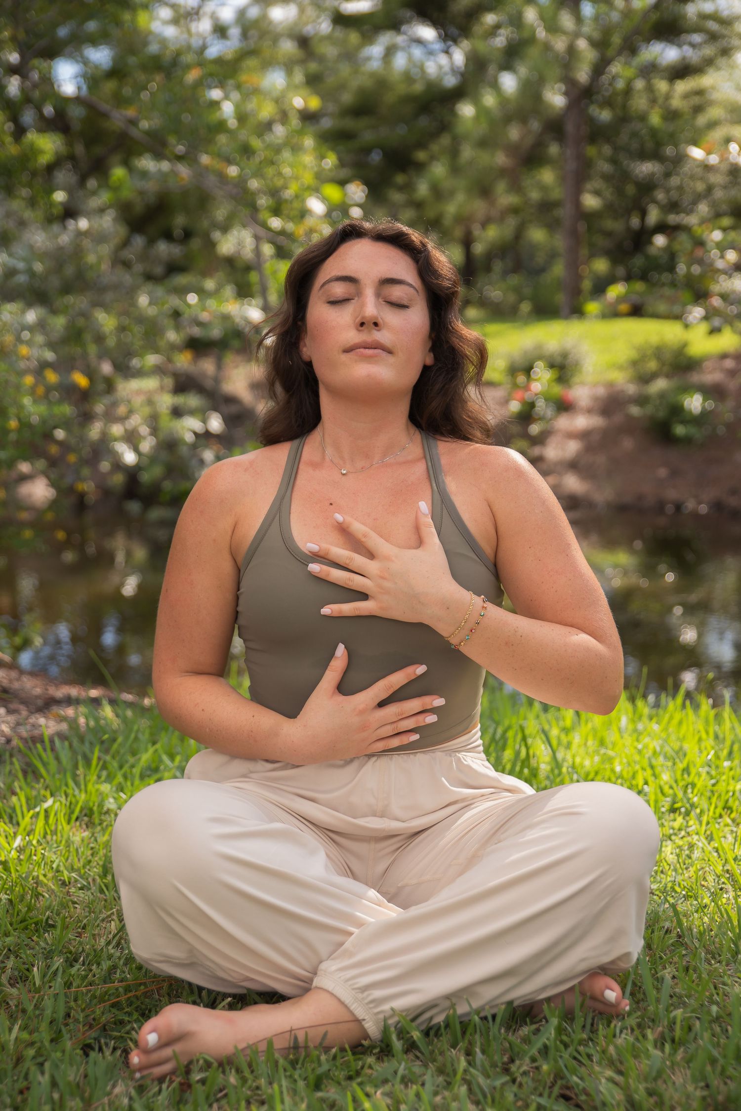 Person in casual attire sitting cross-legged on grass near water with eyes closed in peaceful pose.