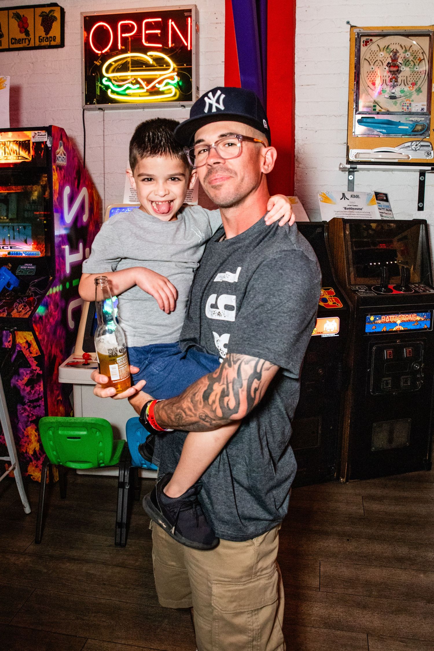 Man with tattoos and NY cap holds smiling boy in an arcade bar with neon open sign.