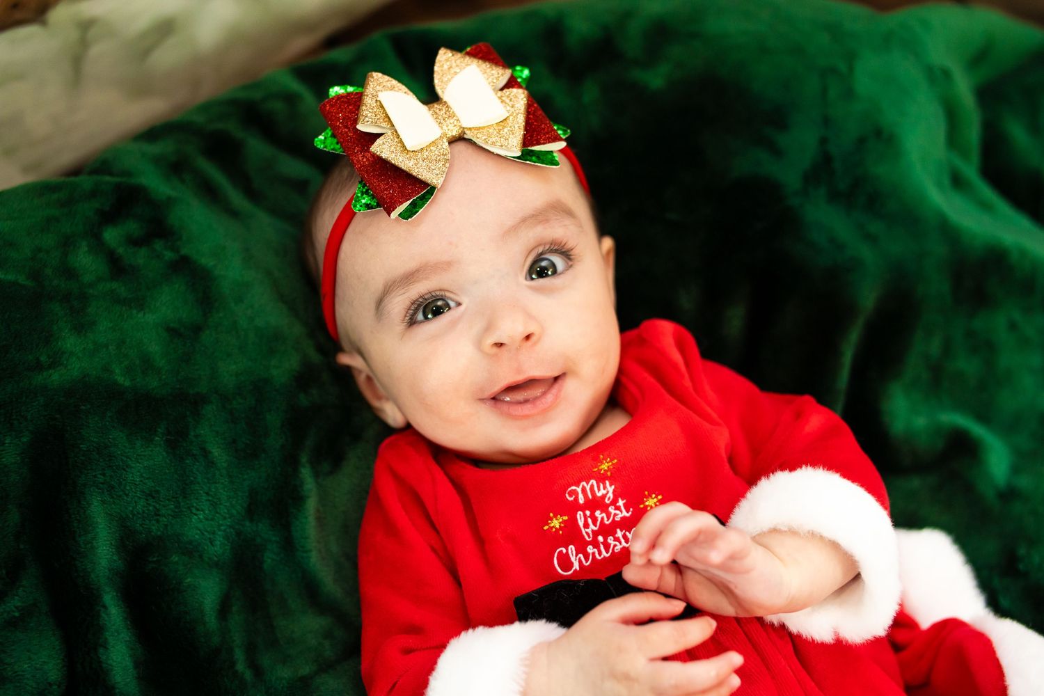 Baby wearing red holiday outfit and bow headband smiles on green velvet background.