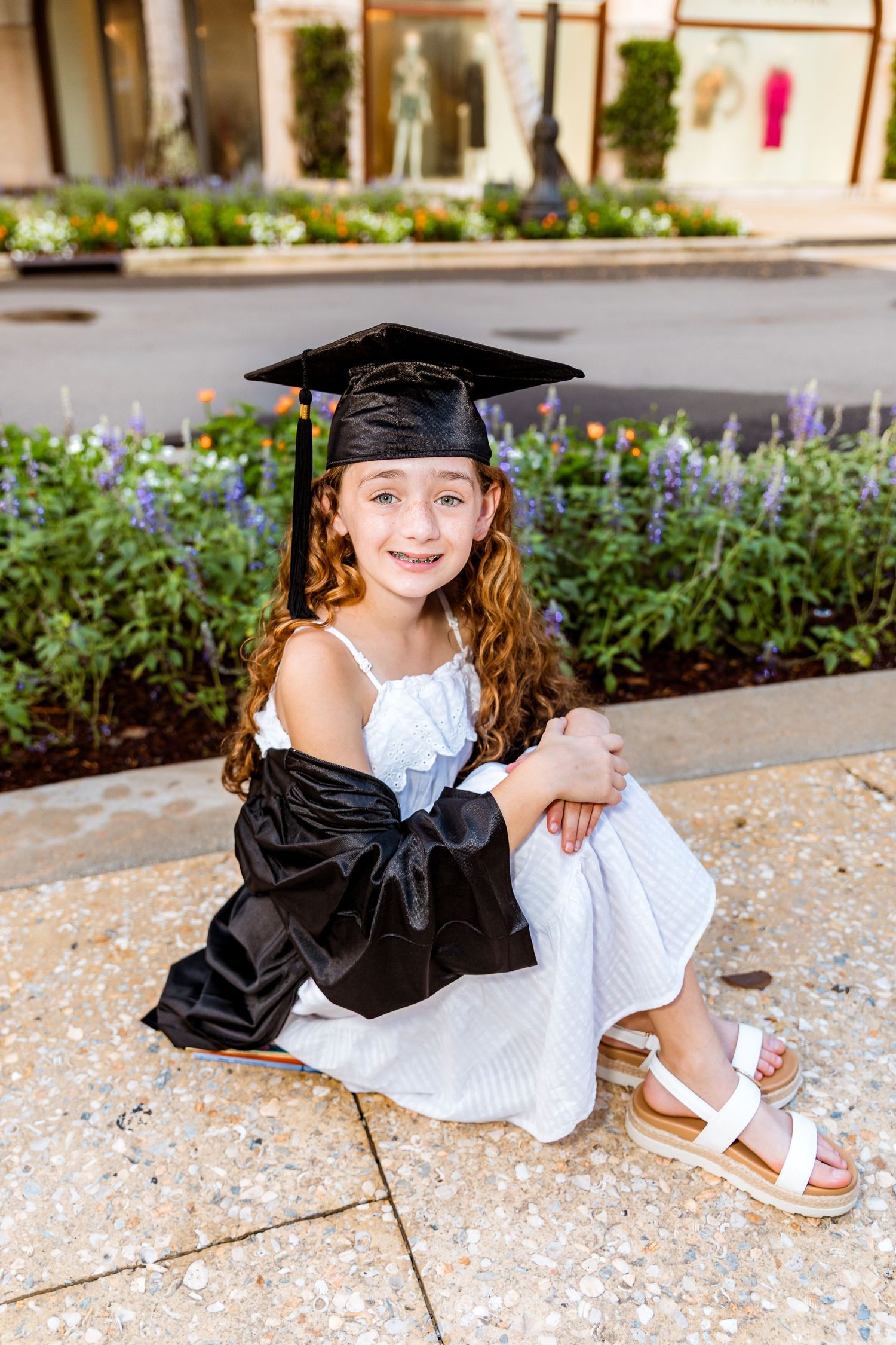 Young girl in graduation cap and gown smiles while sitting outdoors near colorful flower garden.