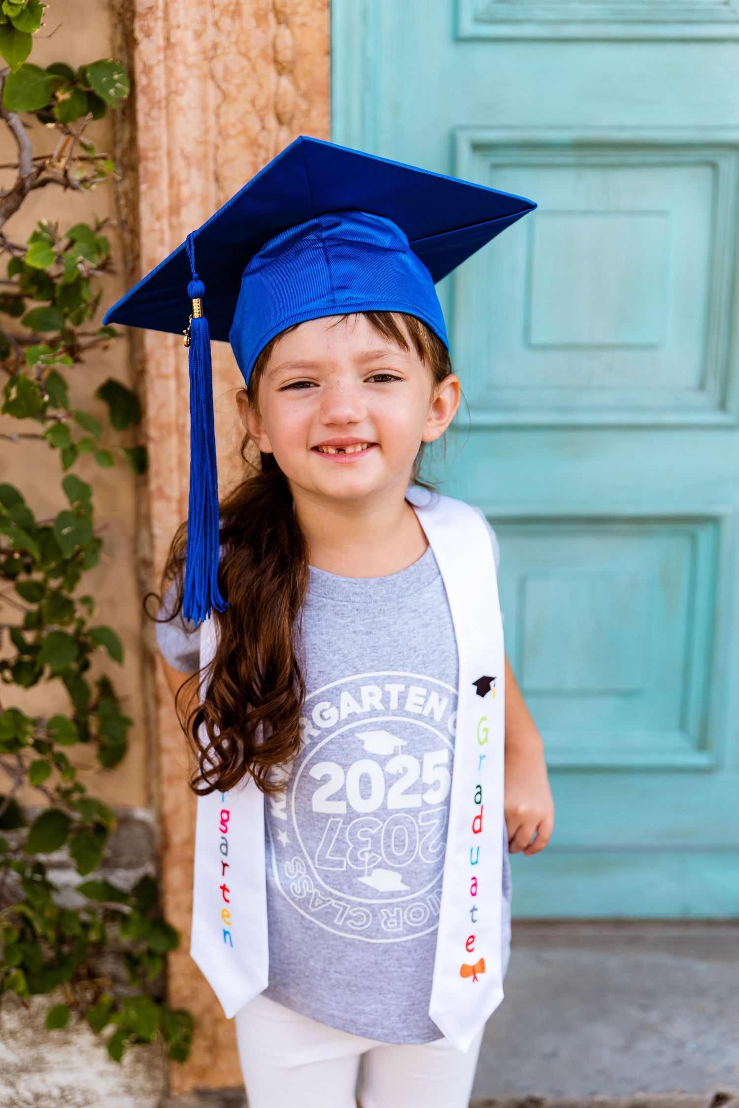 Graduate wearing a blue cap and gown with a 'Class of 2025' shirt poses by a turquoise door with decorative sash.