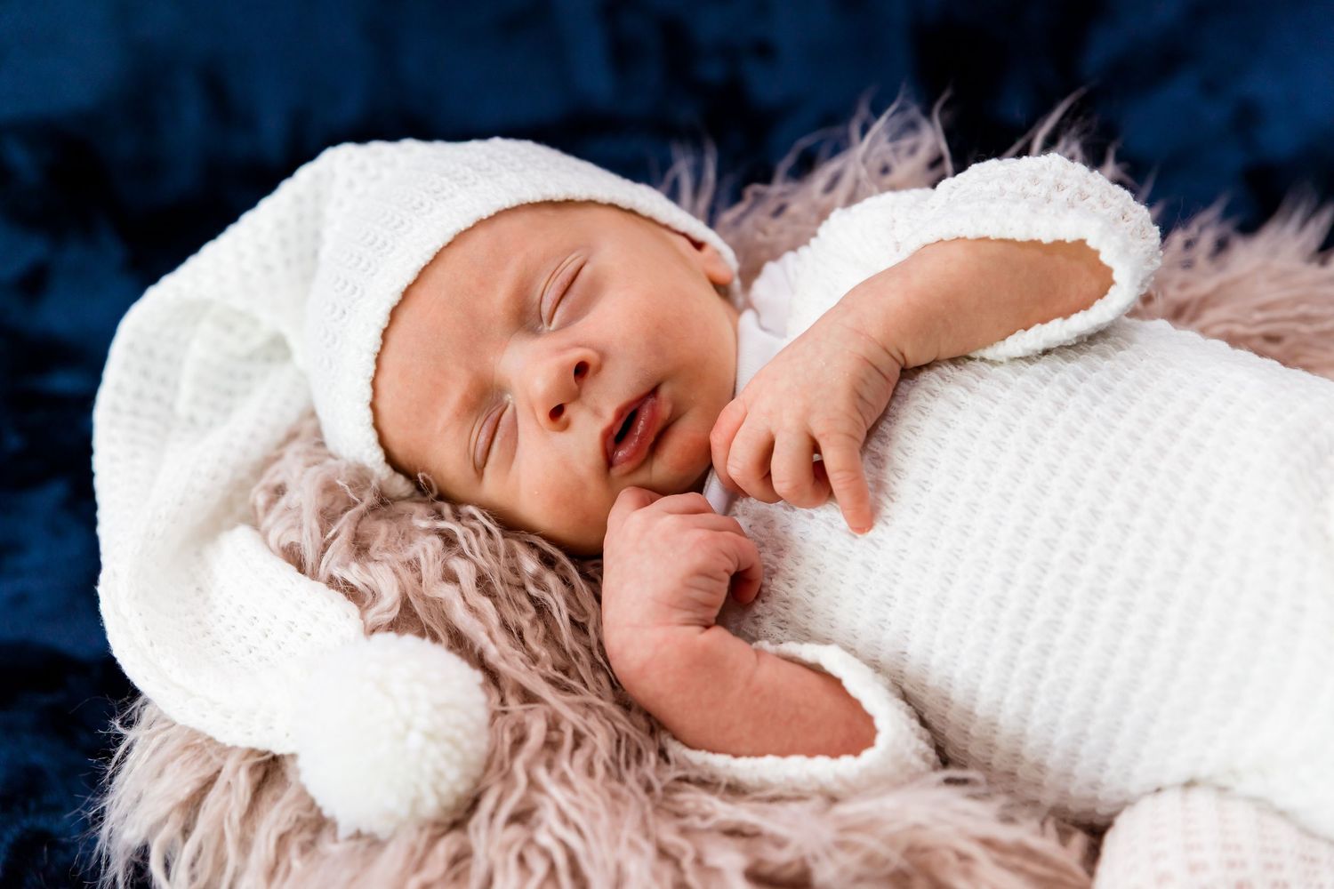 Sleeping newborn in white knit outfit on soft furry blanket against dark blue backdrop.