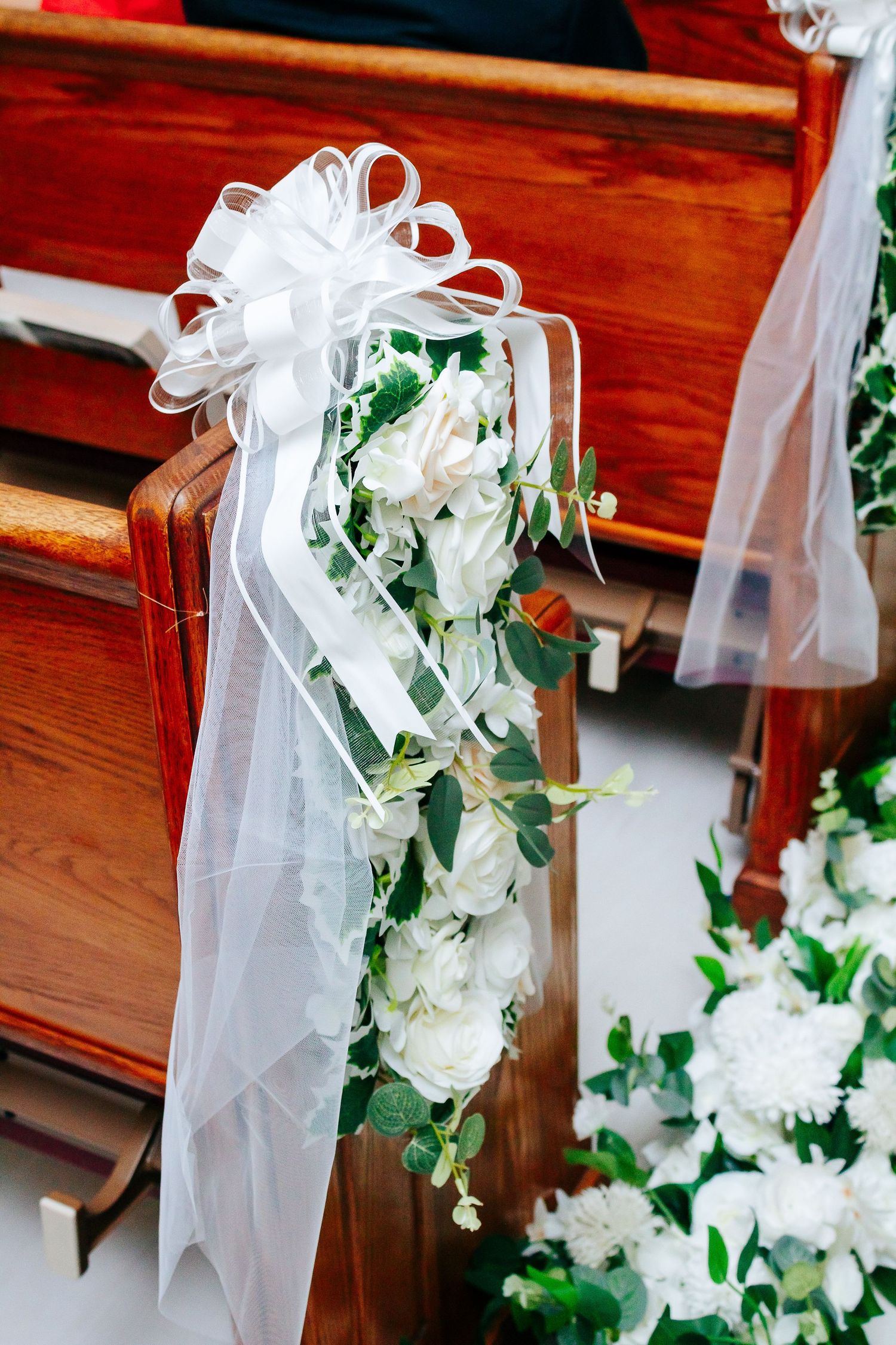 White satin bow and floral church pew decoration with roses and greenery on wooden pew at wedding ceremony.