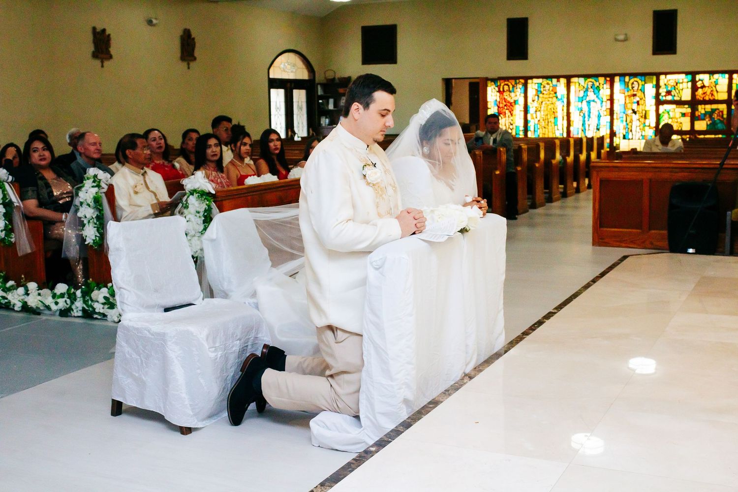 Wedding ceremony taking place in a church with stained glass windows and white-covered chairs.