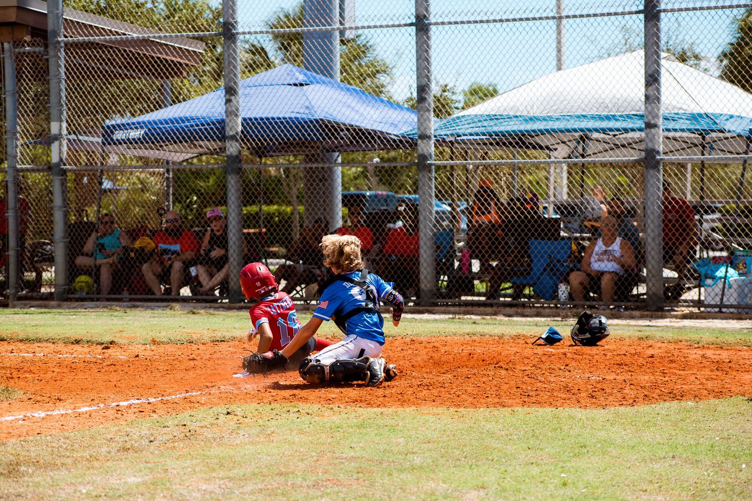 Youth baseball game at field with spectators watching under blue canopy tents.