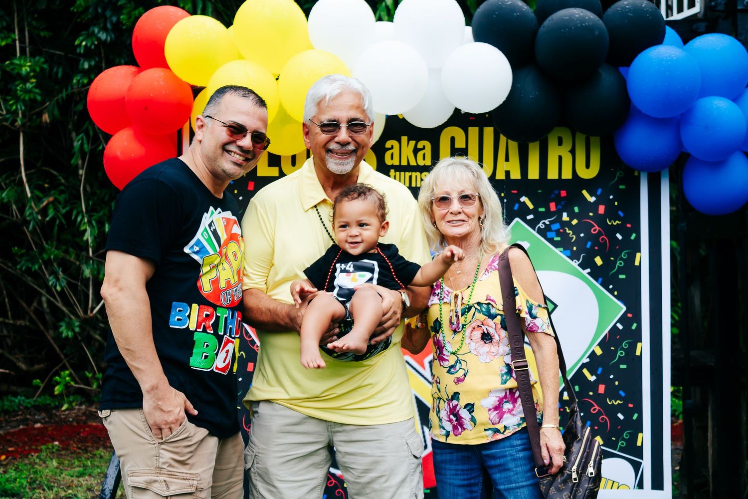 A family celebration outdoors with colorful rainbow balloons and tropical foliage in the background.