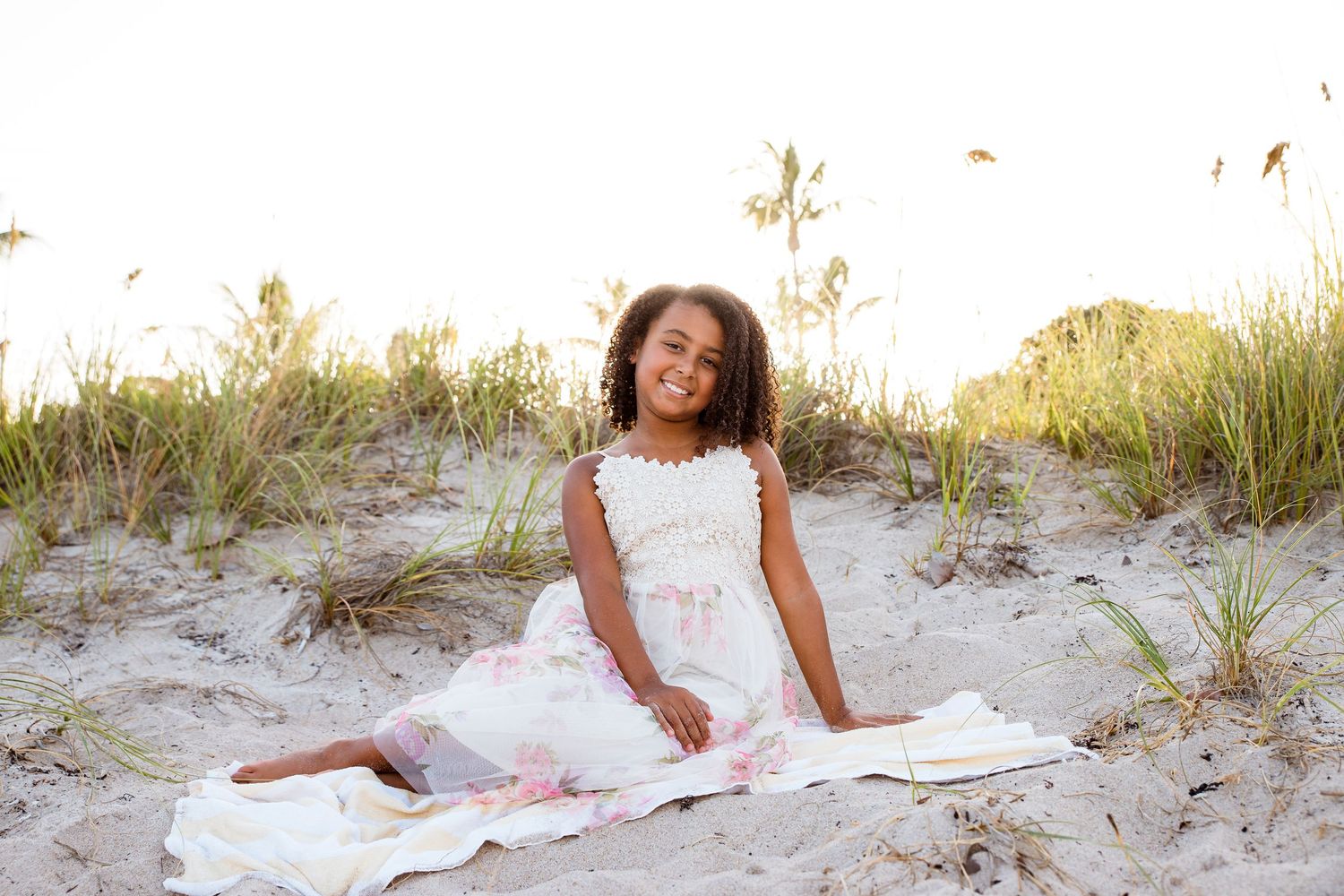A young child in a white dress smiles while sitting on sandy dunes with beach grass in the background at sunset.