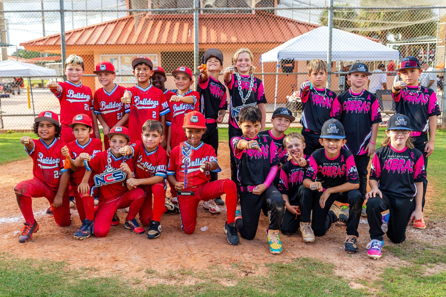 Youth softball team members in pink and black uniforms sitting together on dugout bench for team photo.