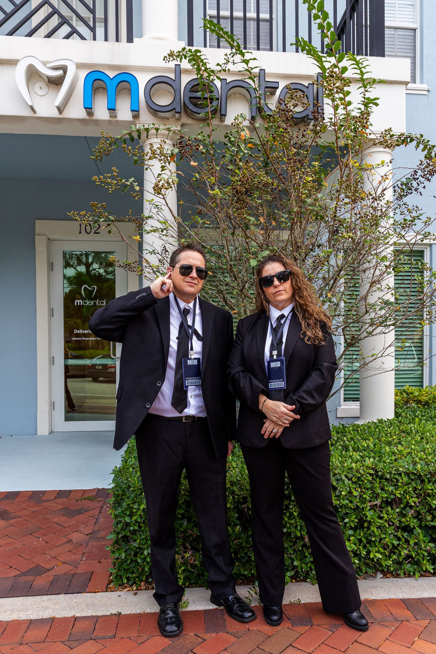 Two people in dark suits and sunglasses pose together on brick pavement outside an office building.