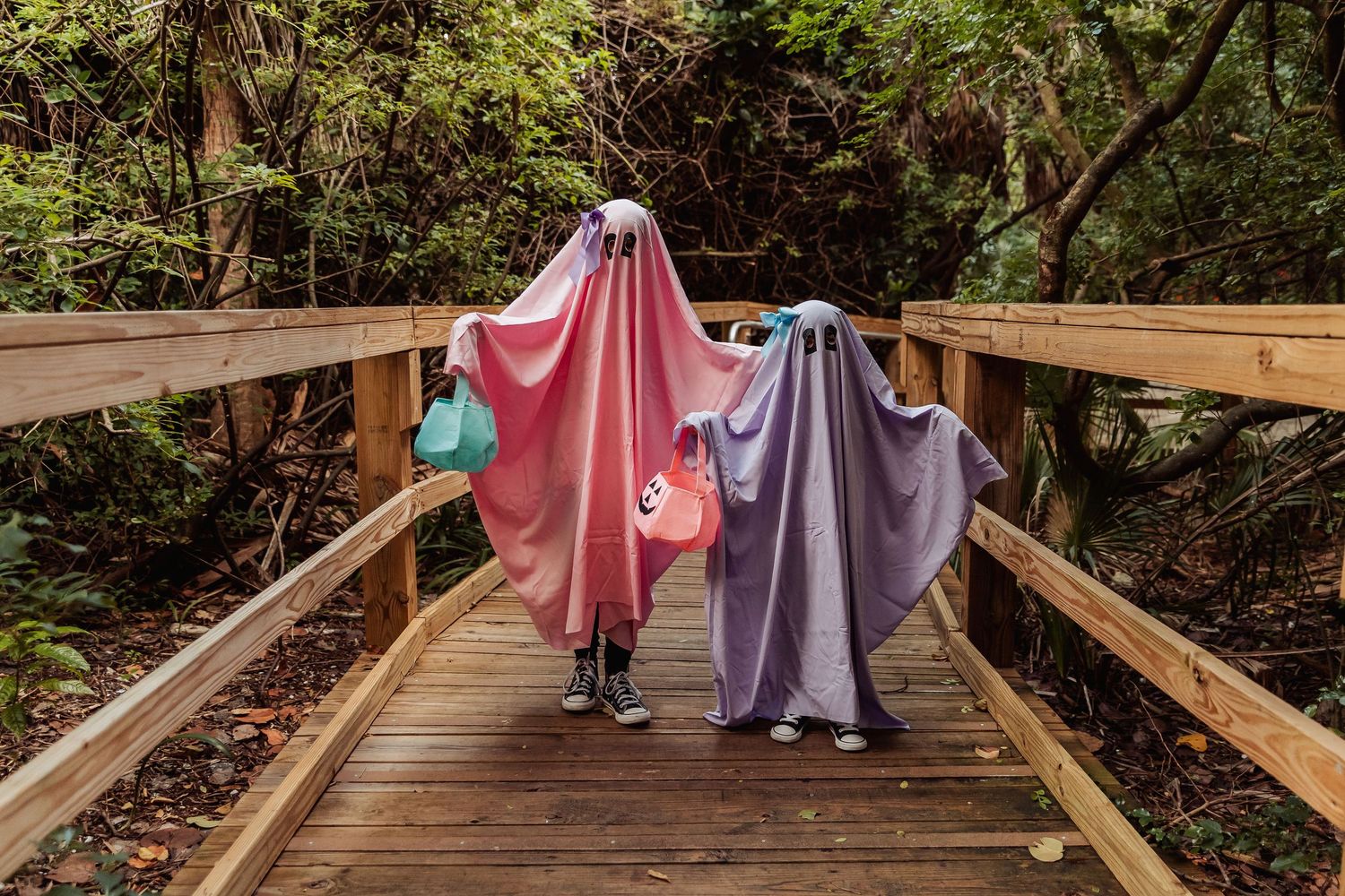 Two people in flowing dresses walk across a wooden bridge in a lush forest setting.
