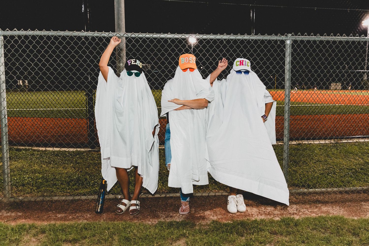 Line of people in white ghost costumes stand along baseball field fence at dusk.