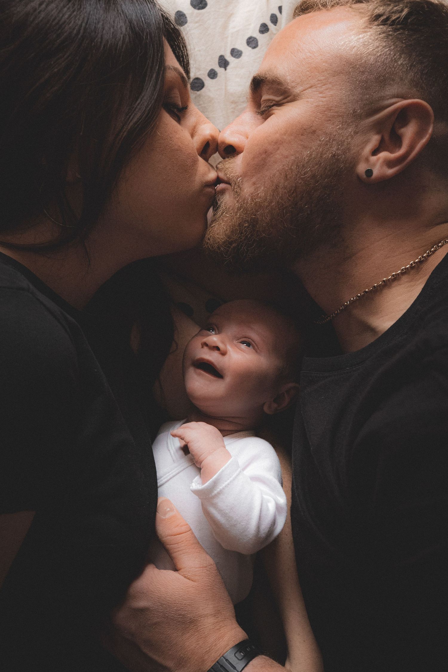 Deux parents qui s'embrassent allongés sur leur lit avec leur bébé de quelques semaines qui les regarde avec un sourire béa