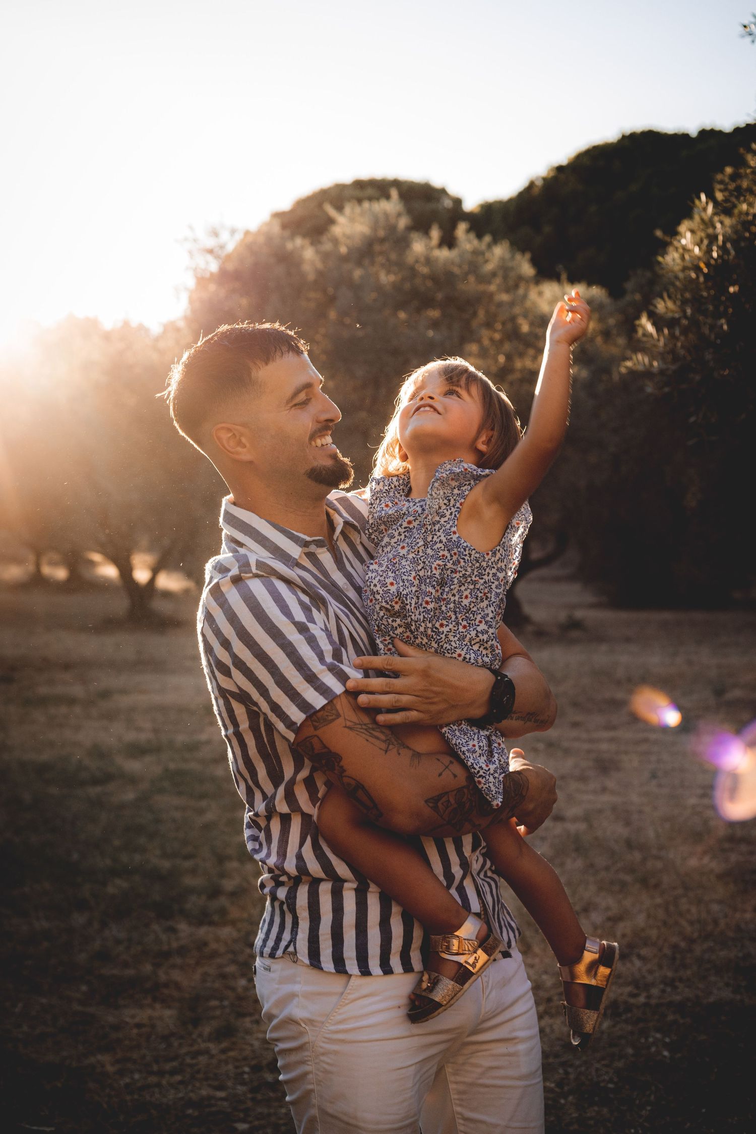 Un jeune papa qui tient sa fille dans ses bras en souriant devant un champ d'olivier en Provence au coucher du soleil d'été