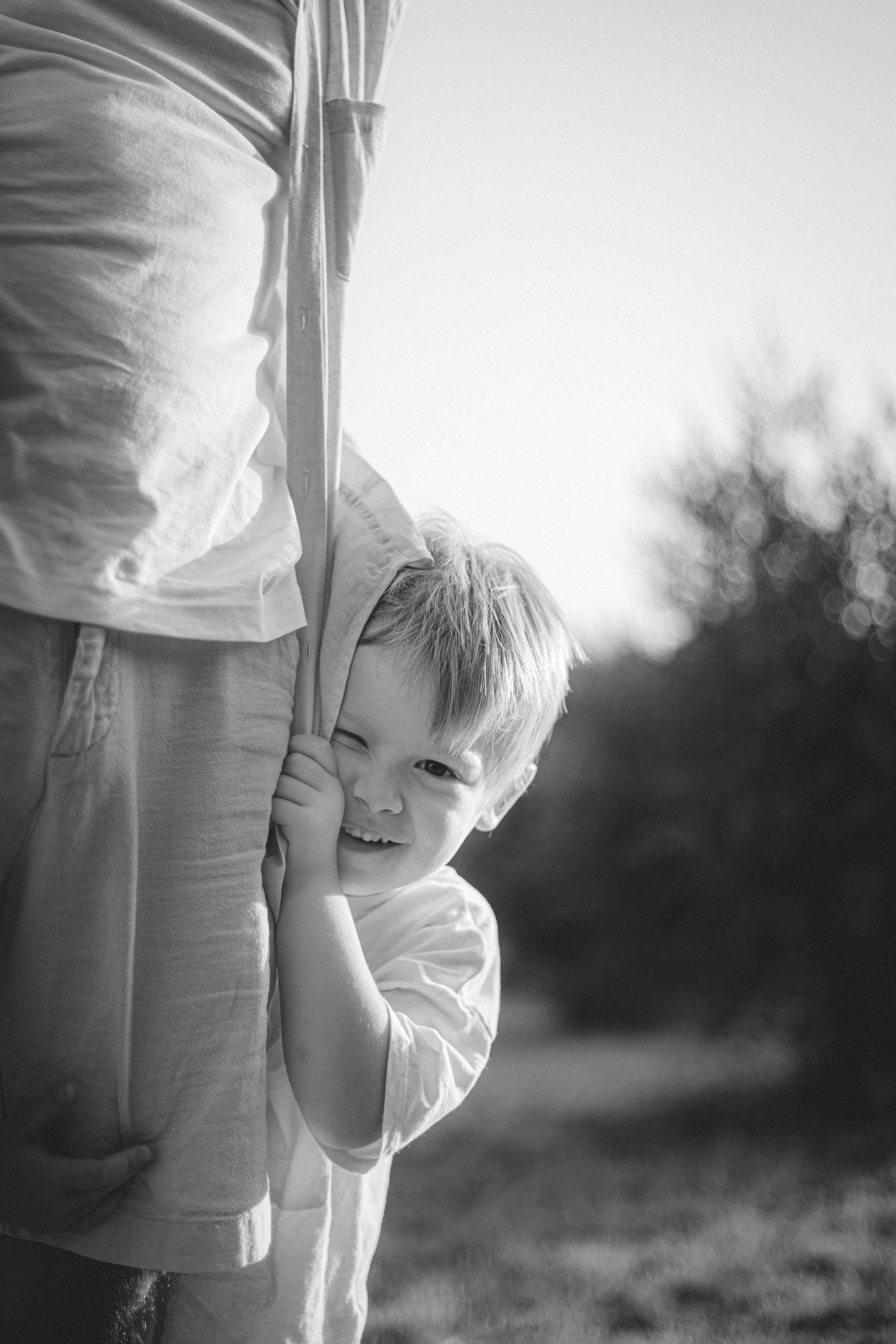 Un jeune enfant au regard malicieux qui fixe l'objectif en se cachant derrière les jambes de son père